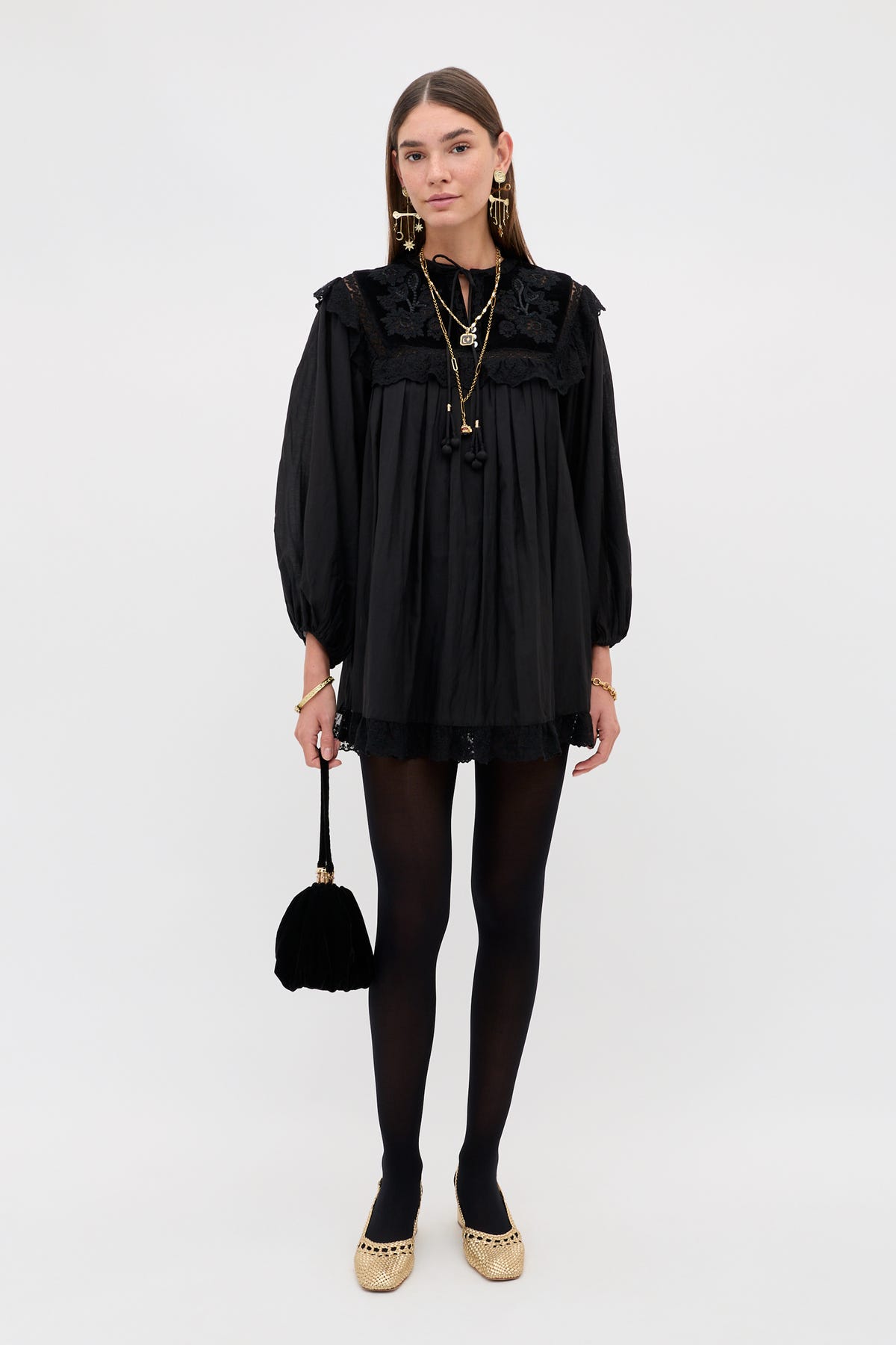 A person wearing the Inez - Lilly Embroidery Black mini dress with long lace sleeves, paired with black tights, metallic flats, and a small black handbag.