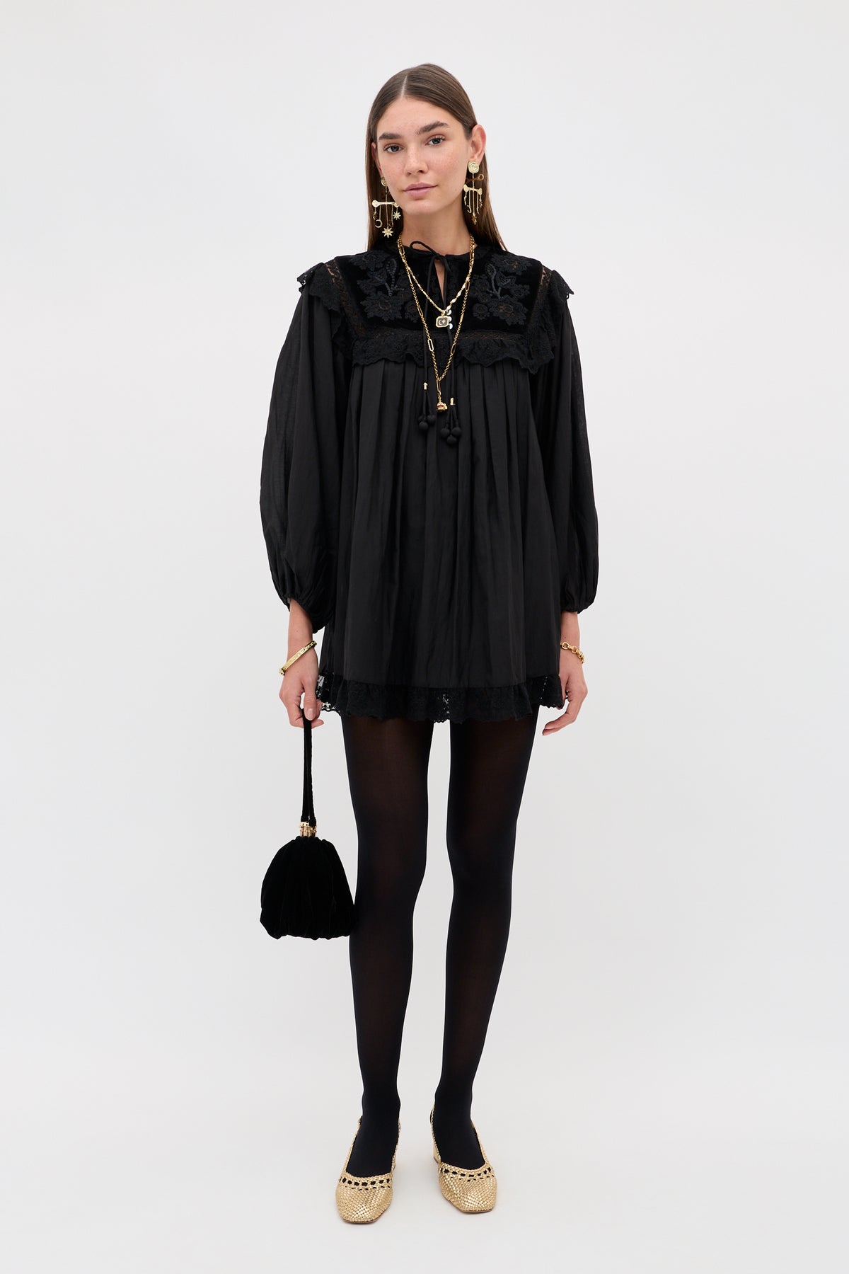 A person wearing the Inez - Lilly Embroidery Black mini dress with long lace sleeves, paired with black tights, metallic flats, and a small black handbag.