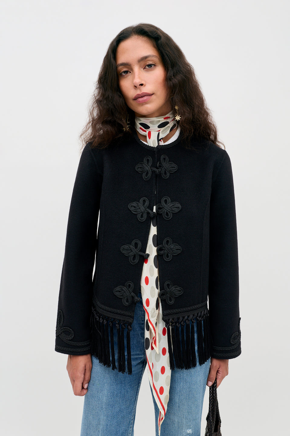 A person with wavy dark hair wears the Inga - Black jacket, featuring ornate closures, along with a red polka dot scarf and blue jeans, standing against a plain white background.