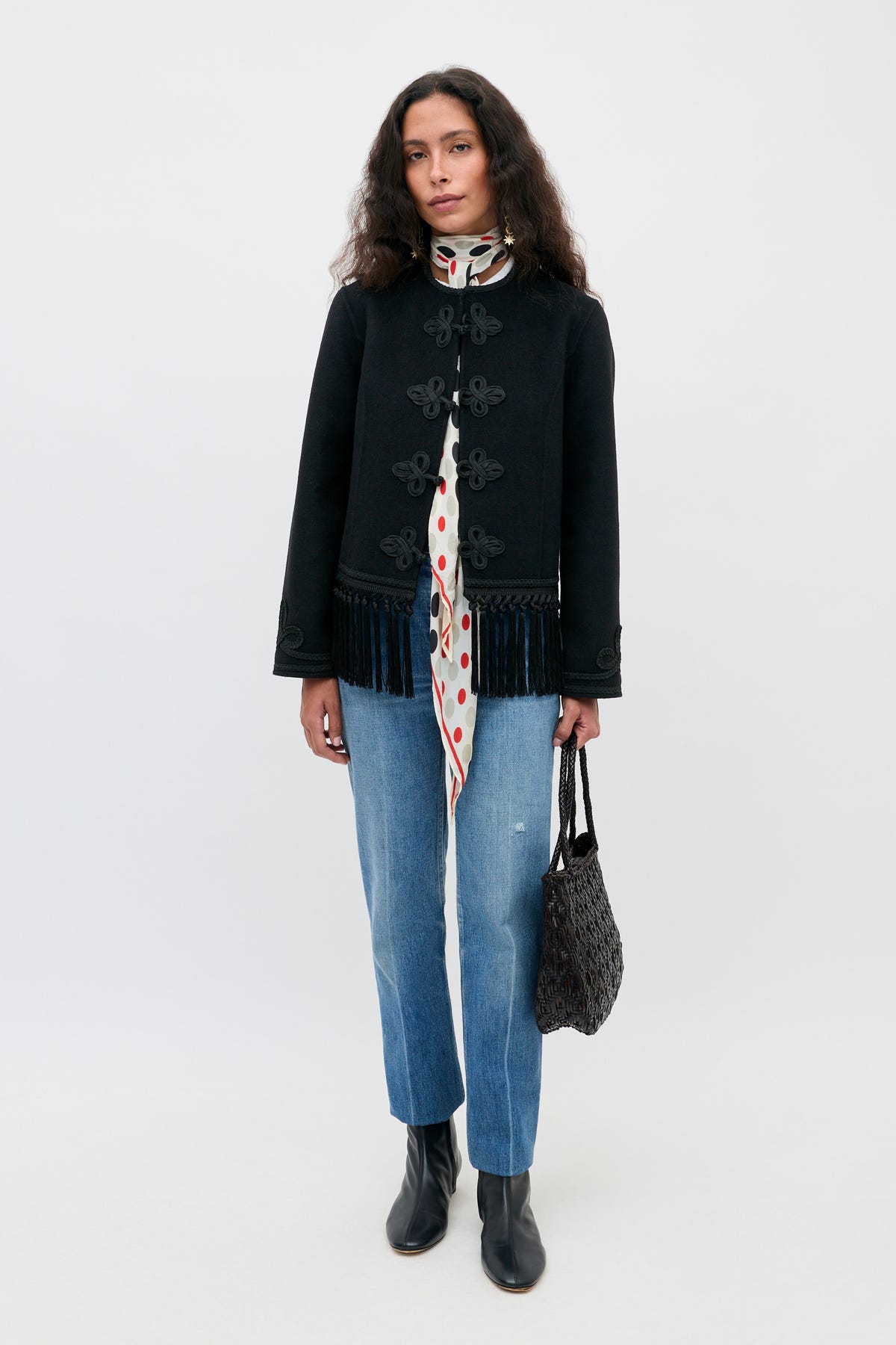 Woman stands against a plain background wearing a black jacket with fringe, a white scarf with red polka dots, blue jeans, black ankle boots, and holding the Inga - Black handbag.