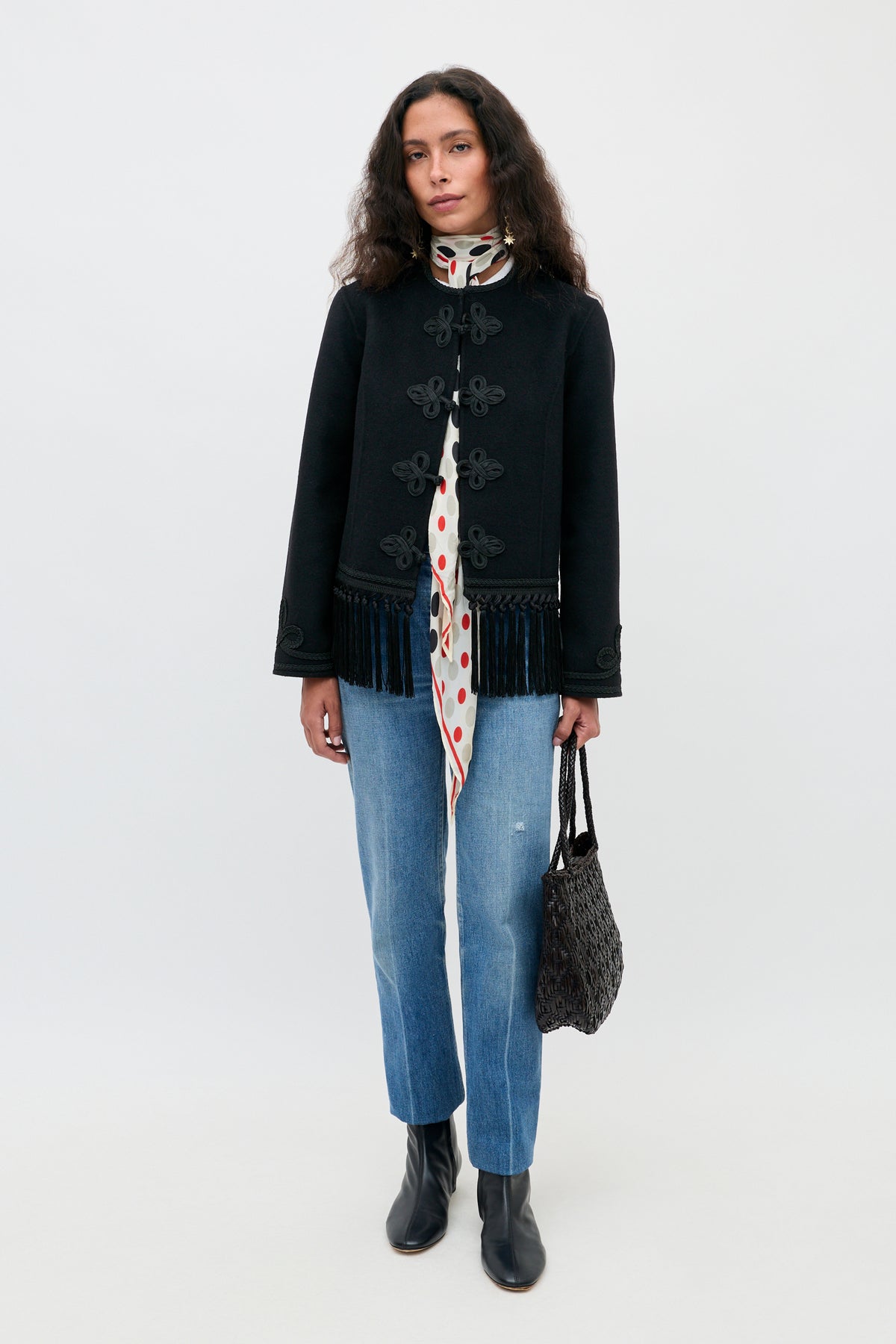 Woman stands against a plain background wearing a black jacket with fringe, a white scarf with red polka dots, blue jeans, black ankle boots, and holding the Inga - Black handbag.