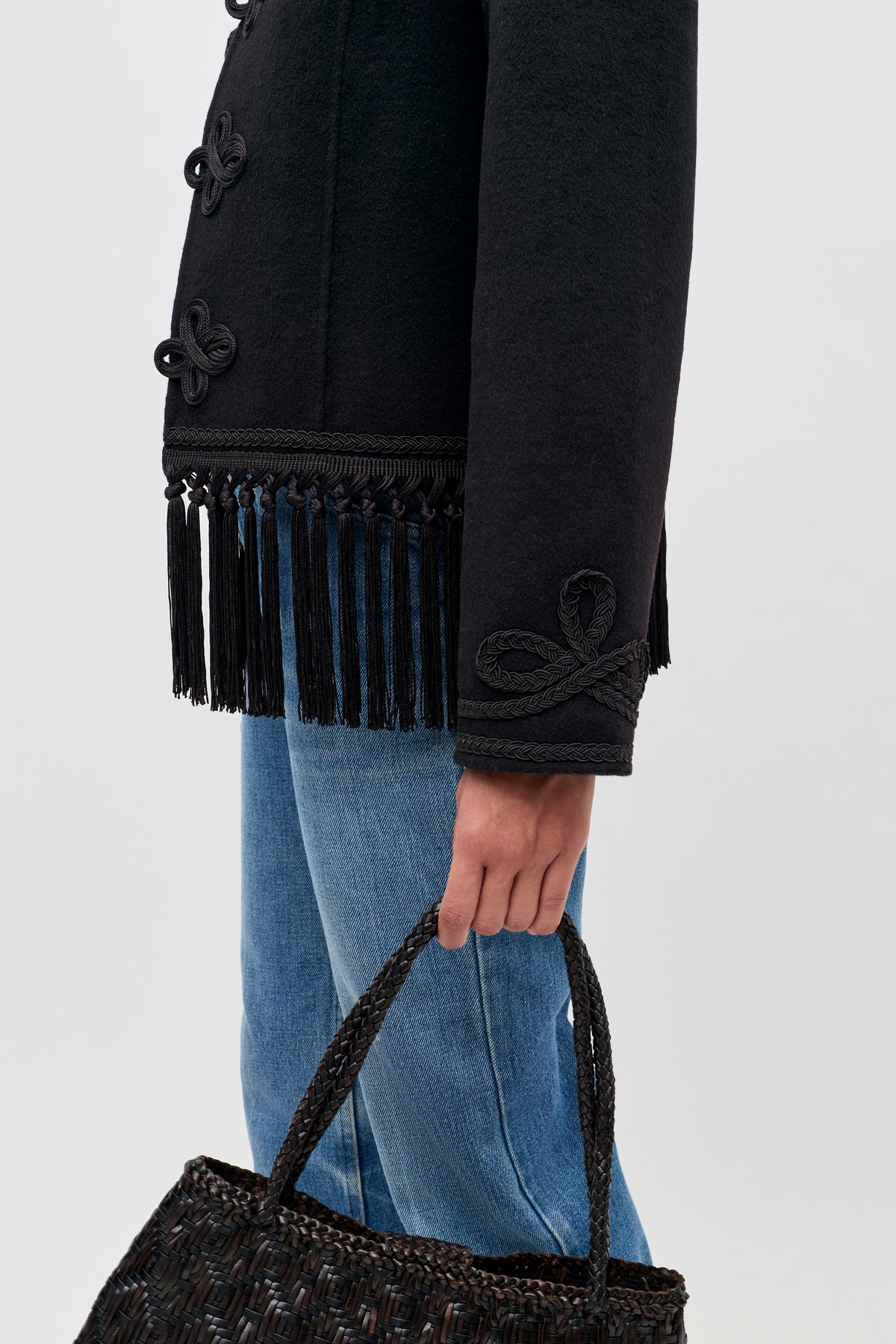 A person wearing the Inga - Black jacket with fringe trim and decorative sleeve details, paired with blue jeans and holding a woven black handbag, stands against a plain background.