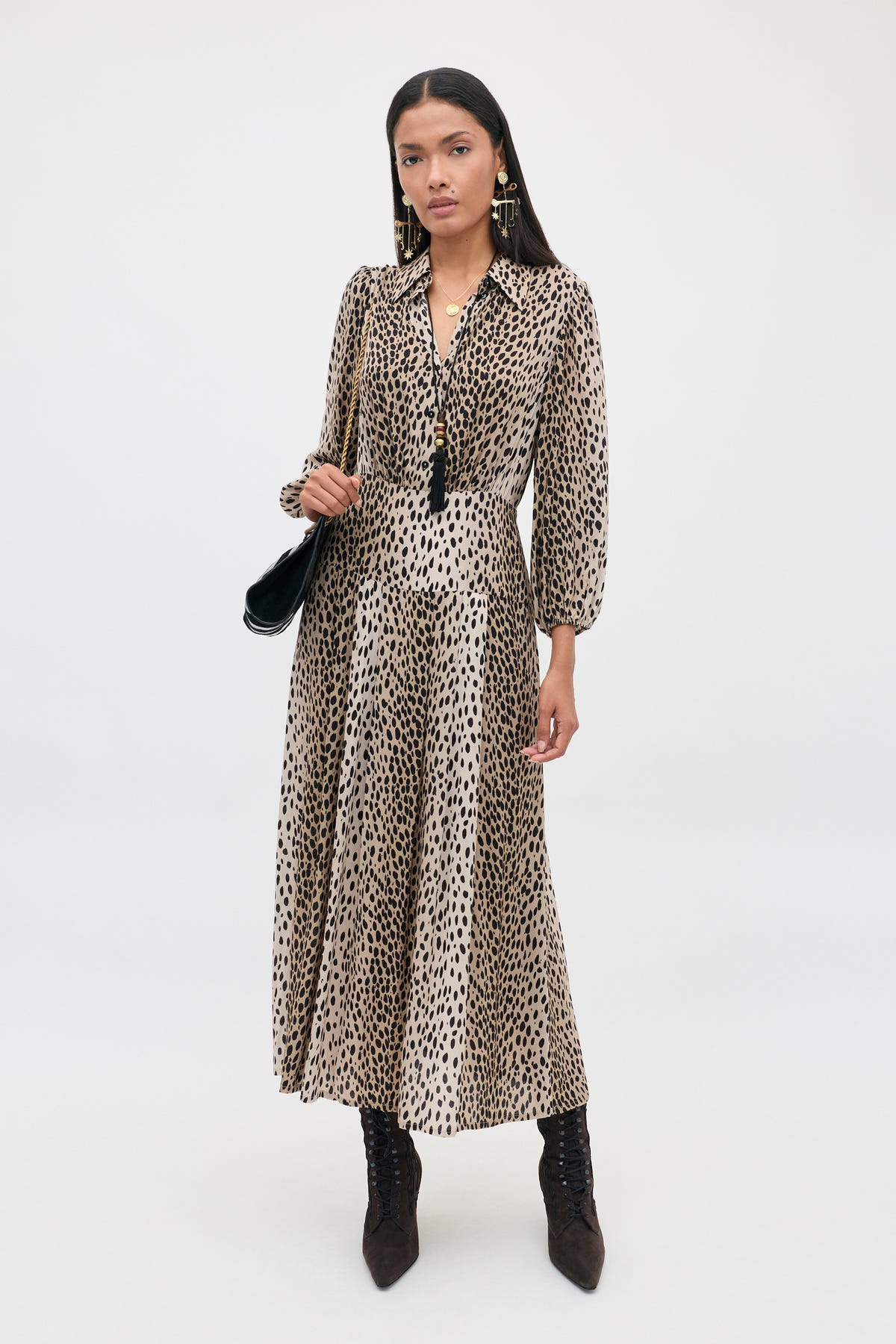 A woman stands against a plain white background, wearing the Izzy - Antelope Spot Black long-sleeved shirt dress with elasticated cuffs, styled with black boots, earrings, a necklace, and holding a black shoulder bag.
