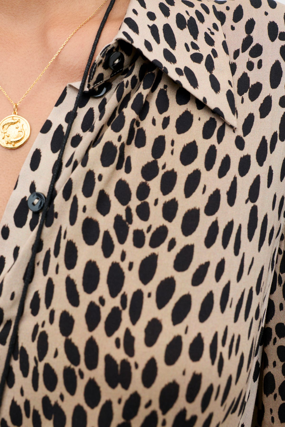 Close-up of the Izzy - Antelope Spot Black blouse, featuring a beige fabric with black spotted pattern, black buttons, elasticated cuffs, and a partially visible gold pendant necklace on the wearer’s neck.