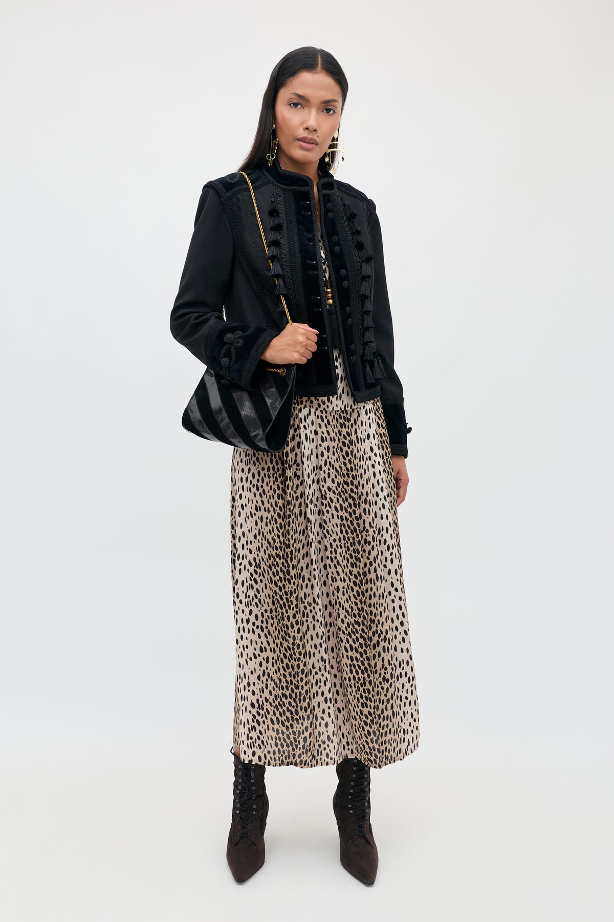 A woman with long dark hair and a neutral expression stands against a plain background, wearing the Izzy - Antelope Spot Black long leopard print skirt, paired with a black jacket featuring elasticated cuffs, black lace-up boots, and holding a black shoulder bag.