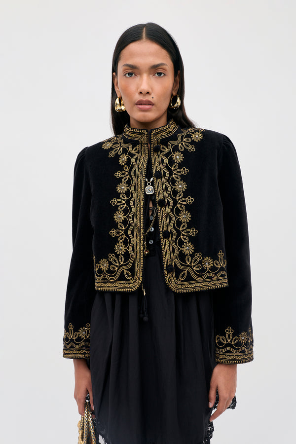 A person wearing the Denver - Soutache Black jacket stands against a light grey background, looking directly at the camera. The black cotton velvet jacket features intricate gold embroidery that adds an elegant touch to their refined appearance. They have long dark hair and wear gold earrings.