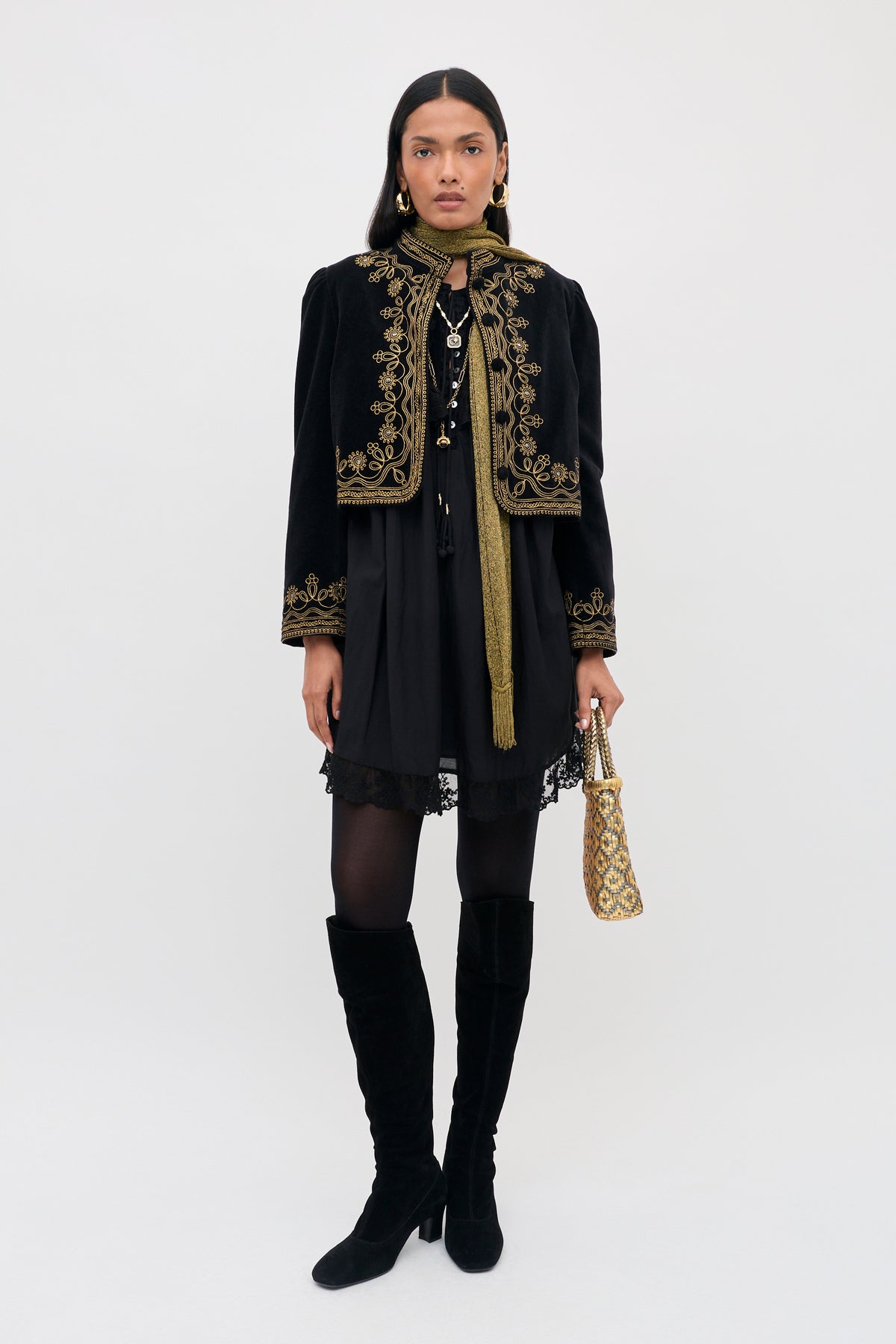 A woman stands against a plain light background wearing the Denver - Soutache Black: a striking black velvet jacket adorned with gold embroidery, styled with a black dress with lace trim, black tights, knee-high black boots, a gold scarf, and a patterned gold handbag.