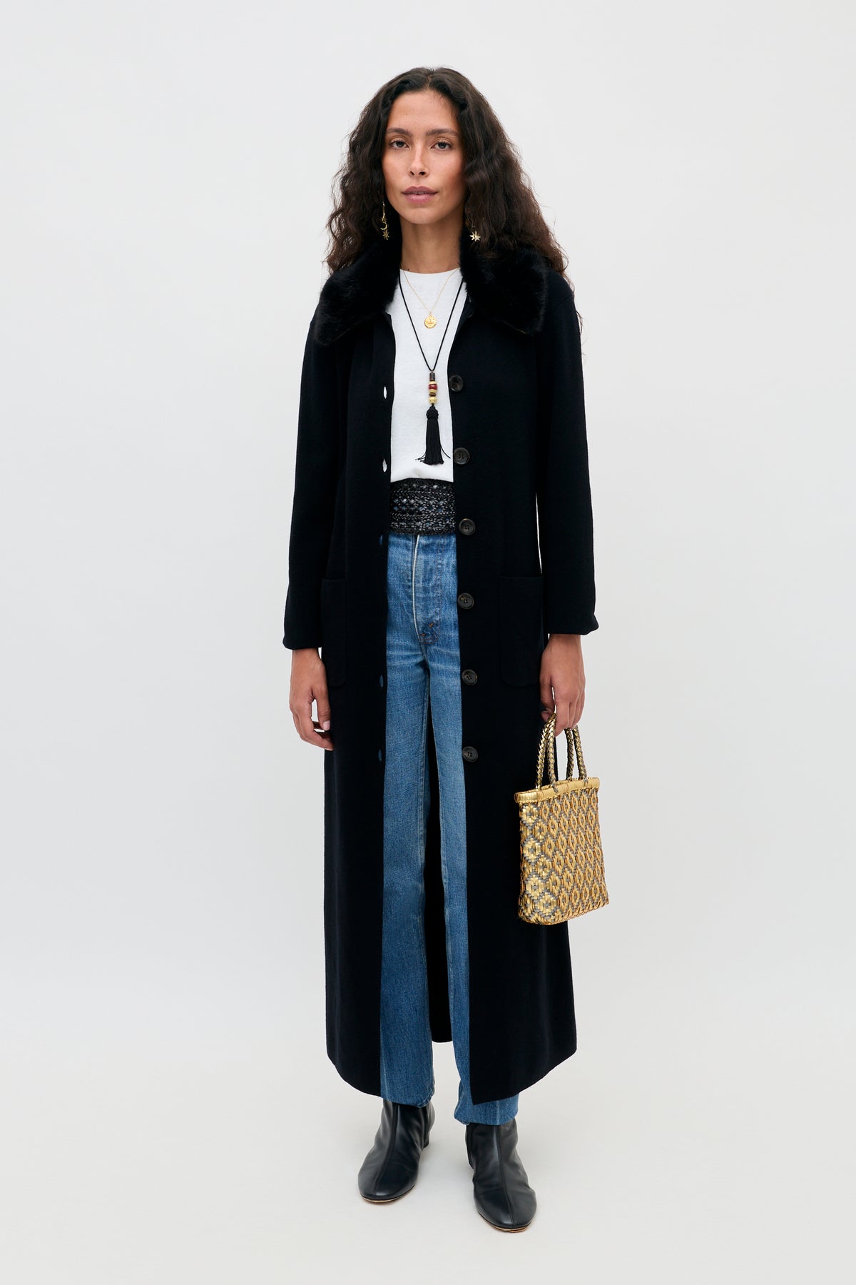 A woman wearing a Jacoba - Black coat with a faux fur collar over a white t-shirt, blue jeans, and black boots, holding a woven gold-tone handbag.