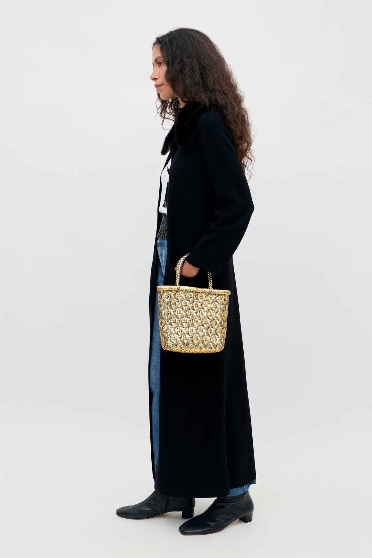 A woman in a long black coat and jeans carries a small metallic woven Jacoba - Black handbag.