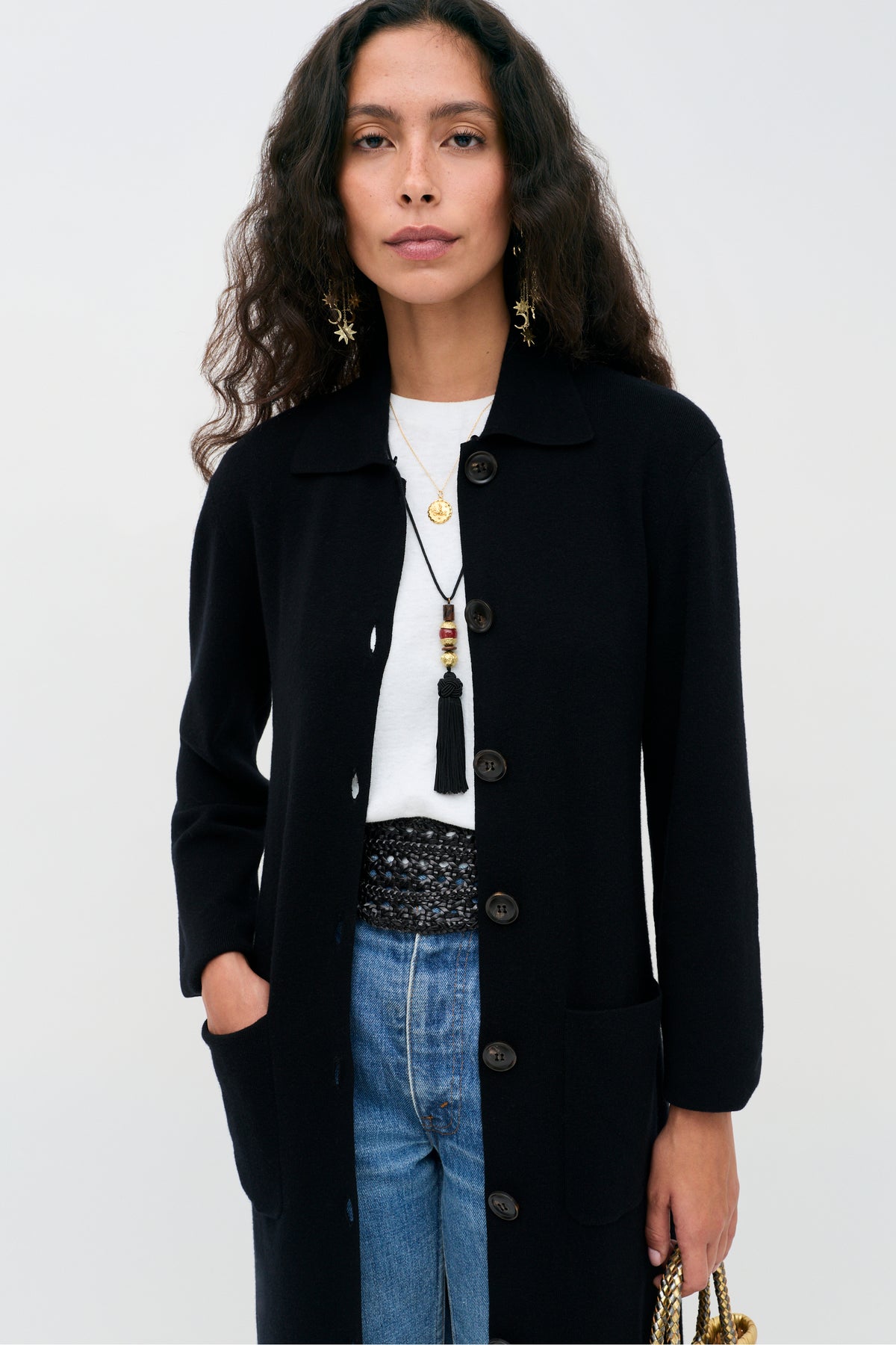 A person wearing a Jacoba - Black long-sleeved button-down cardigan over a white t-shirt and blue jeans, accessorized with layered gold necklaces and star-shaped earrings.