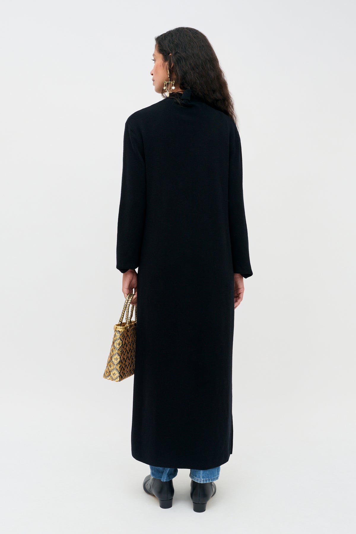 A person stands facing away wearing a Jacoba - Black long-sleeved coat over jeans and black ankle boots, holding a small patterned handbag.