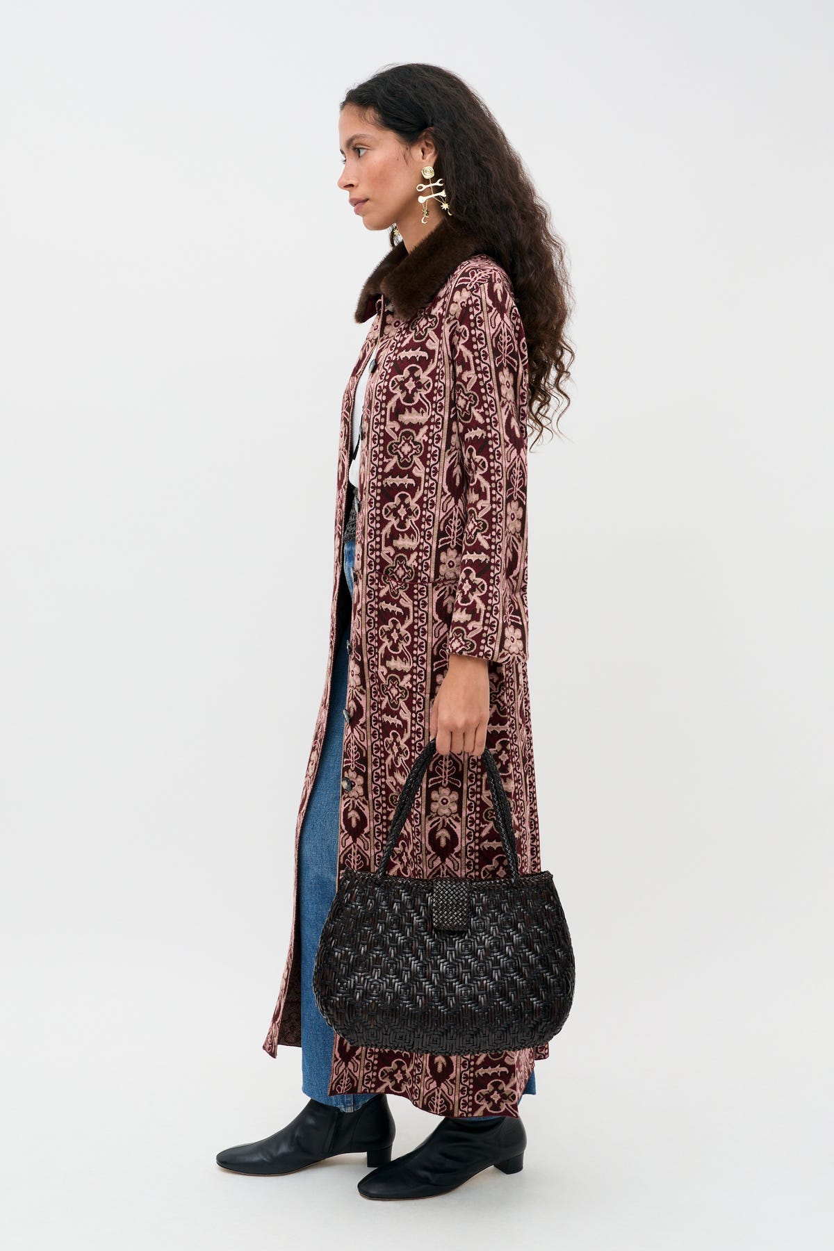 A woman stands in profile wearing the Jacoba - Tapestry Bloom Bordeaux, a long patterned coat, paired with blue jeans and black boots. She holds a black textured handbag, accessorizes with large earrings, and wears her long dark curly hair down. The background is plain white.
