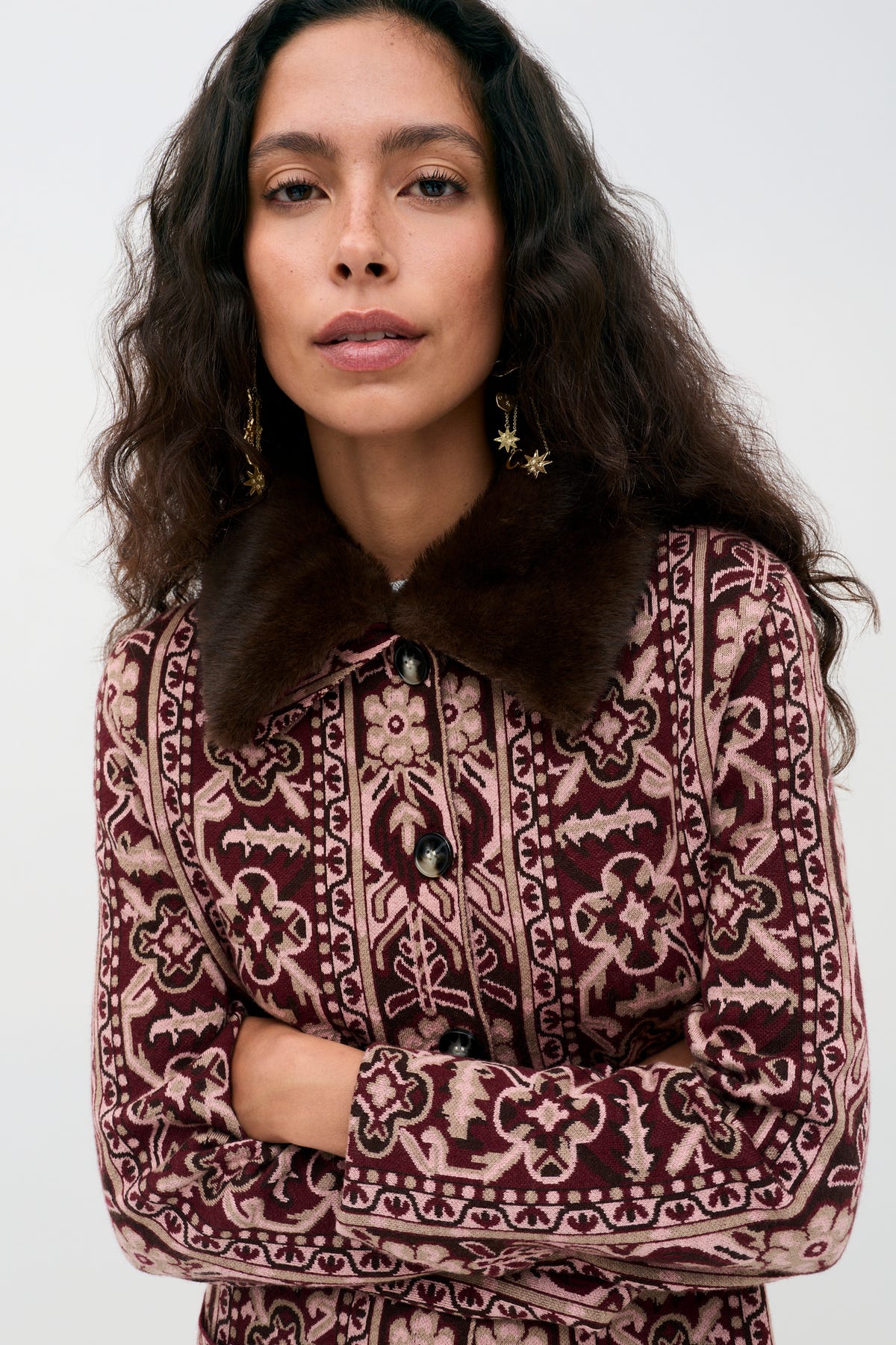 A woman with long, wavy hair wears the Jacoba - Tapestry Bloom Bordeaux coat with a dark fur collar and star-shaped earrings, standing against a plain light background.