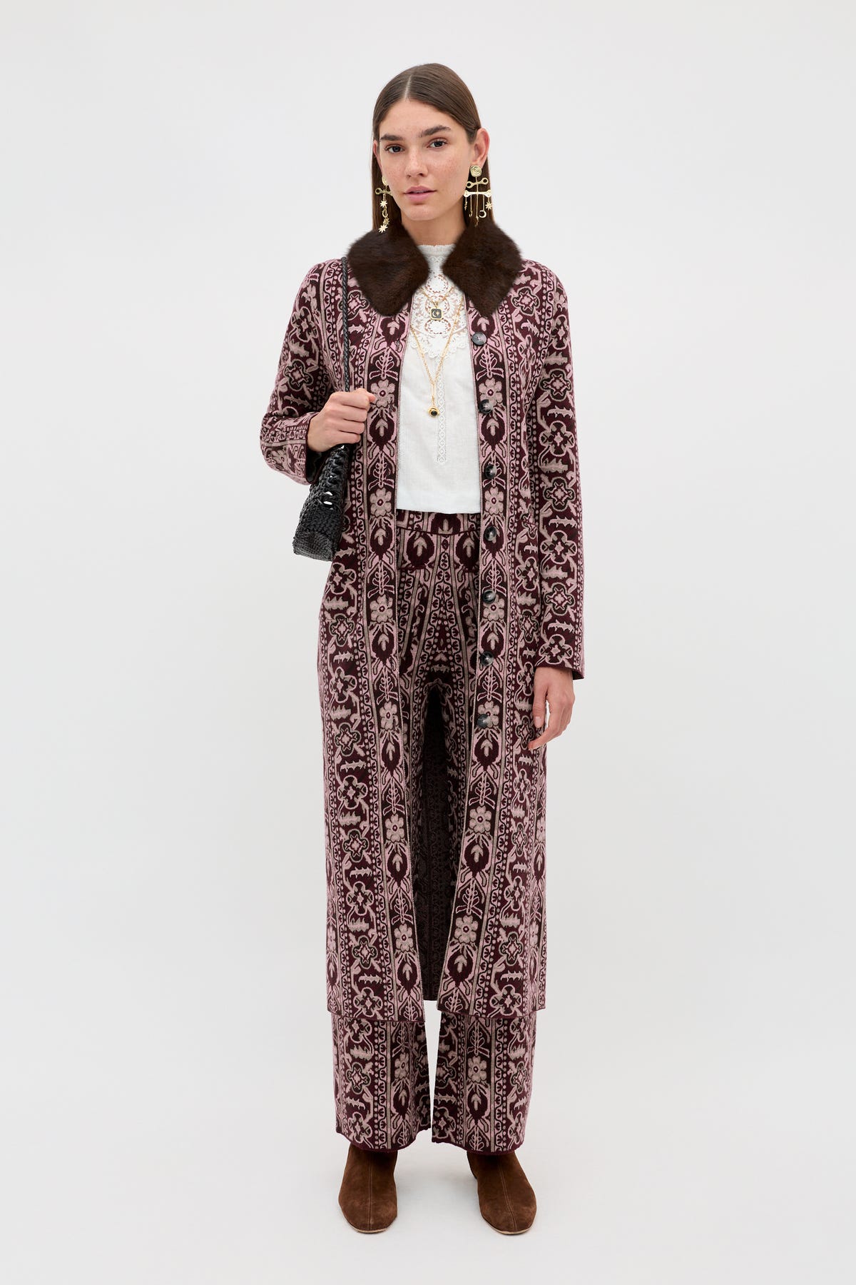 A woman wears the Ceara - Tapestry Bloom Bordeaux long patterned coat with matching pants, paired with a white top, layered necklaces, brown shoes, and a black handbag, standing against a plain white background.