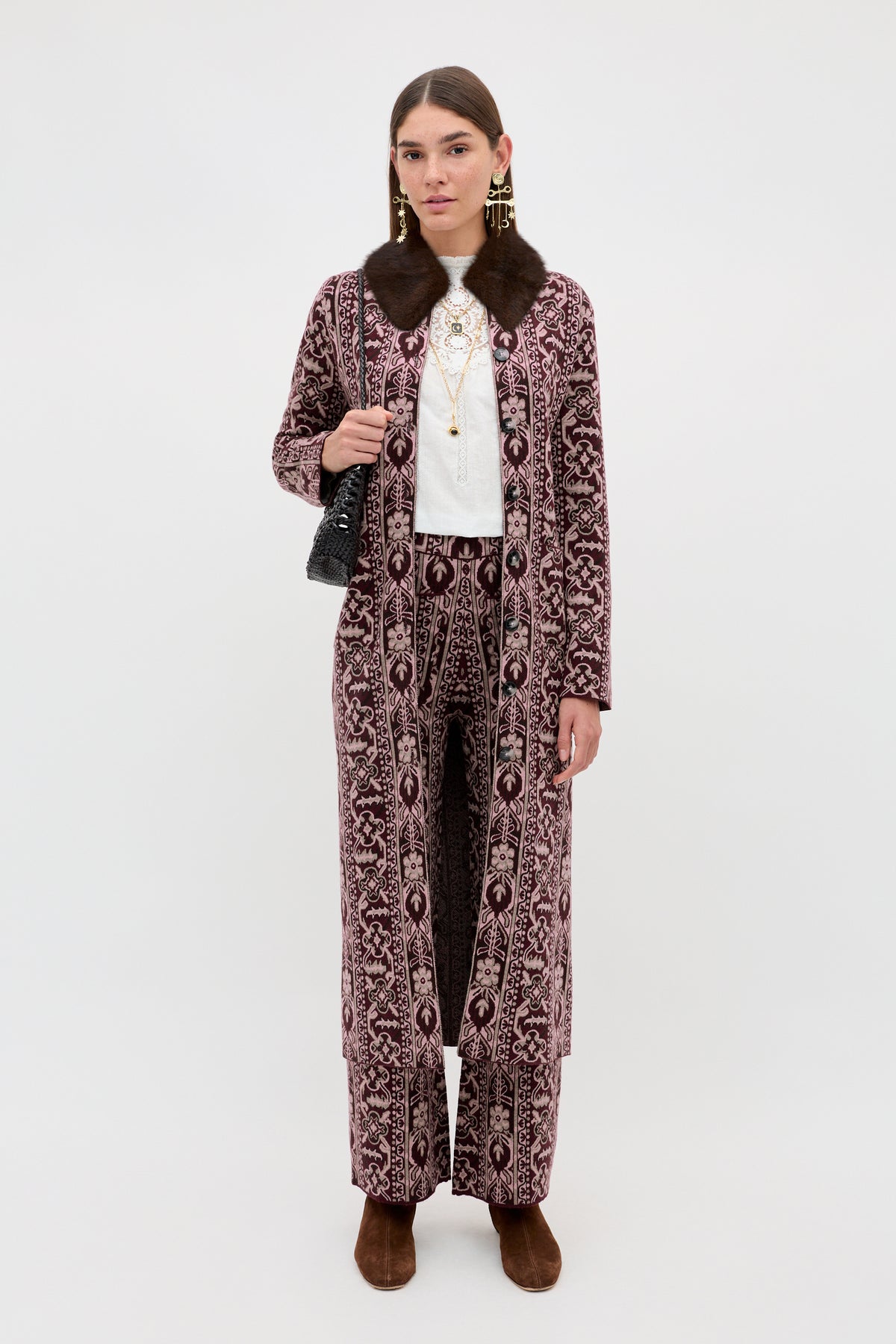 A woman wears the Ceara - Tapestry Bloom Bordeaux long patterned coat with matching pants, paired with a white top, layered necklaces, brown shoes, and a black handbag, standing against a plain white background.