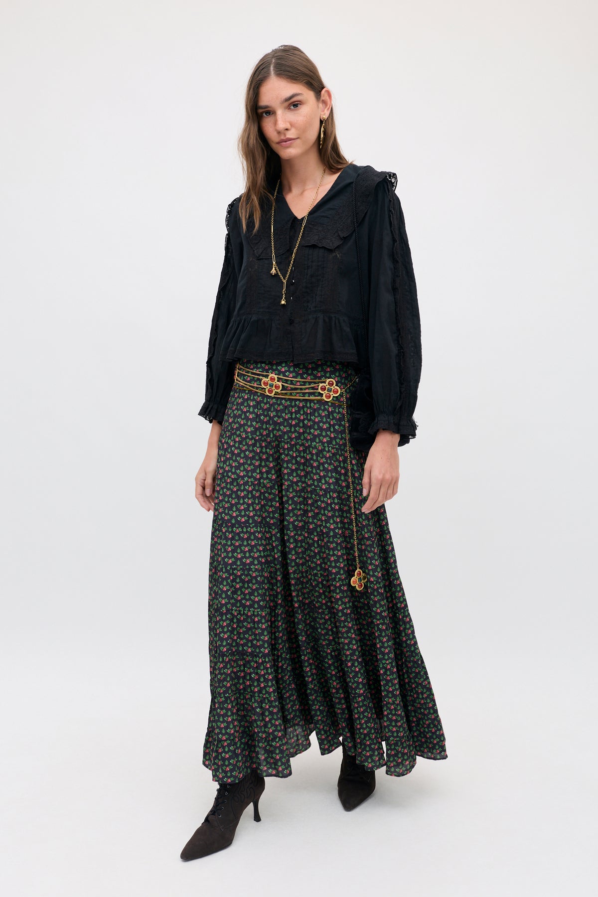 A woman wears a black long-sleeved blouse with a lace collar and a Joey - Mini Rose Pink floral-print maxi skirt cinched by a gold chain belt with flower charms.