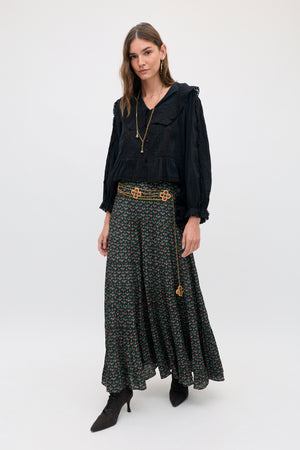 A woman wears a black long-sleeved blouse with a lace collar and a Joey - Mini Rose Pink floral-print maxi skirt cinched by a gold chain belt with flower charms.