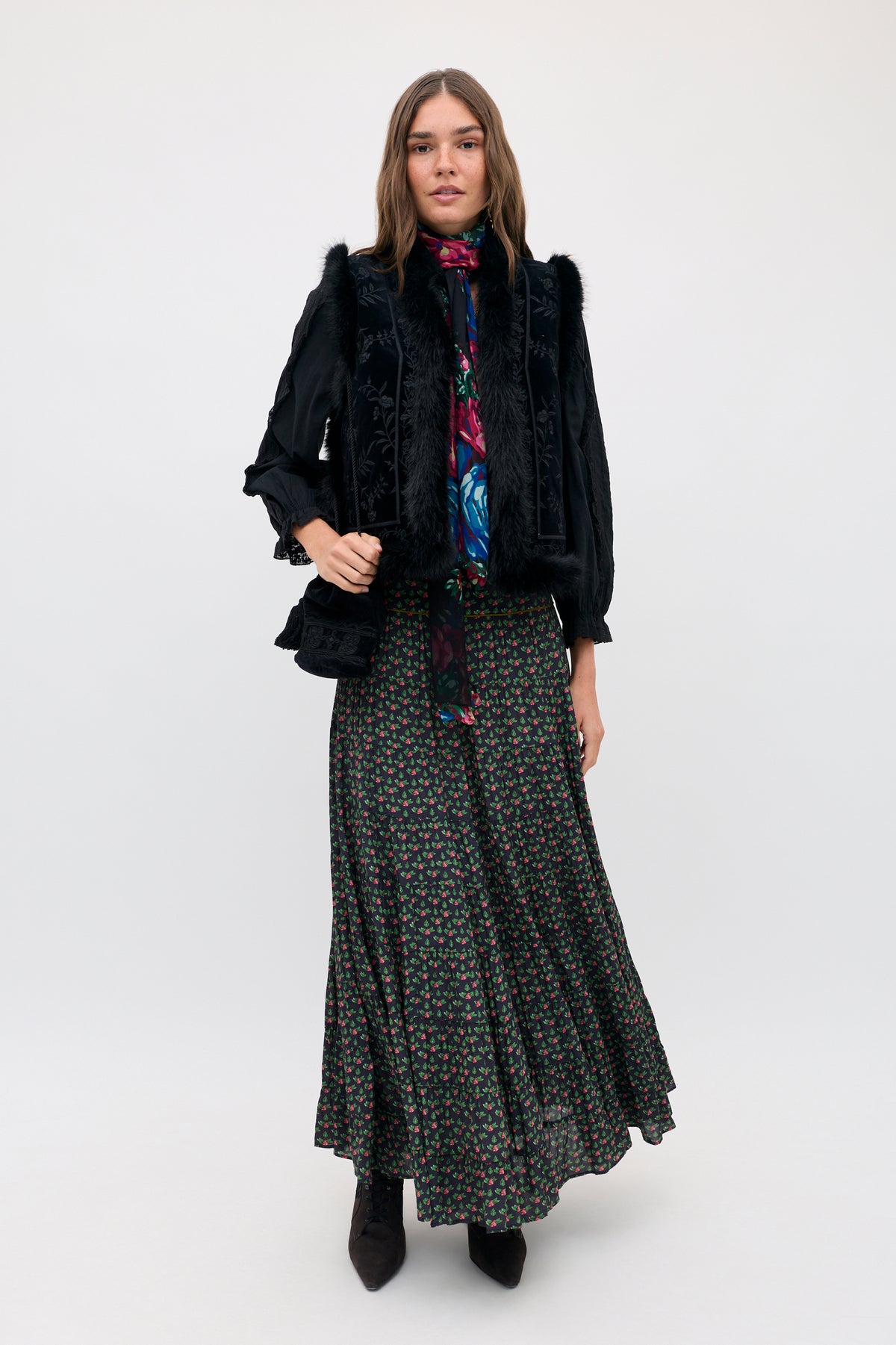 A woman wearing a Joey - Mini Rose Pink black floral maxi skirt, a dark patterned blouse with a neck tie, and a black vest with faux fur trim.