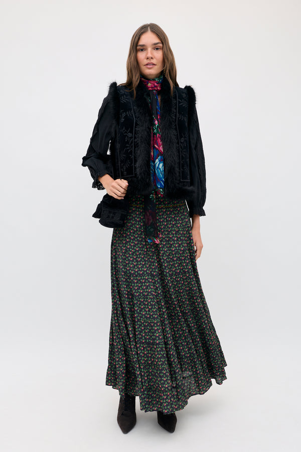 A woman wearing a Joey - Mini Rose Pink black floral maxi skirt, a dark patterned blouse with a neck tie, and a black vest with faux fur trim.