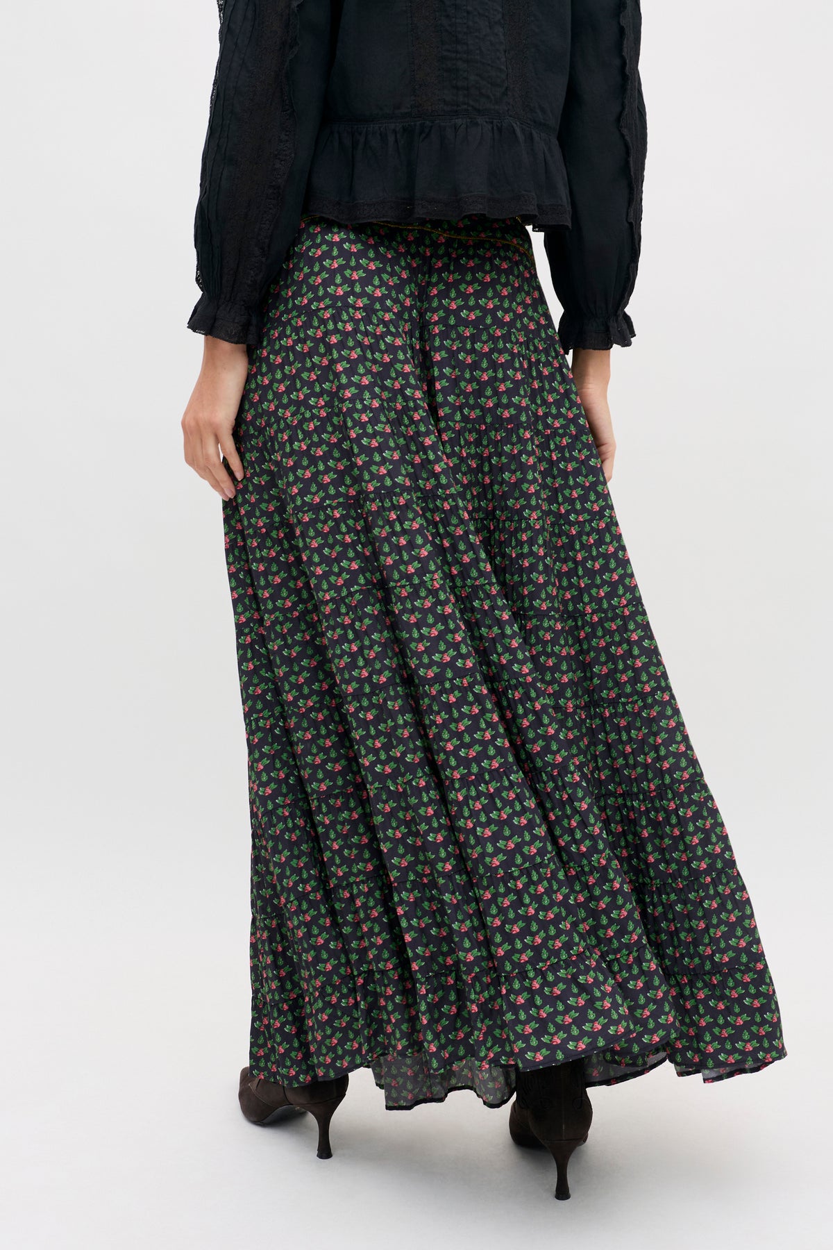 A person wears a black, long-sleeved blouse tucked into a Joey - Mini Rose Pink tiered maxi skirt featuring a small pink rose floral pattern.