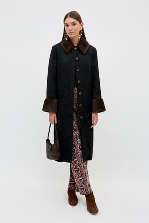 A woman wearing the Jonah - Black long coat with a brown fur collar and cuffs, styled with patterned pants, brown boots, and a small textured brown handbag, stands against a plain white background.