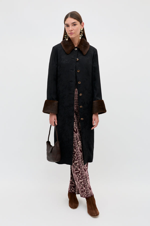 A woman wearing the Jonah - Black long coat with a brown fur collar and cuffs, styled with patterned pants, brown boots, and a small textured brown handbag, stands against a plain white background.