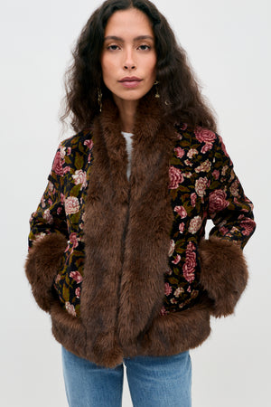 A woman wearing the Jorgia - Enchanted Flora Green jacket, featuring a floral pattern and brown faux fur trim on the collar, cuffs, and hem, paired with blue jeans and standing against a plain light background.