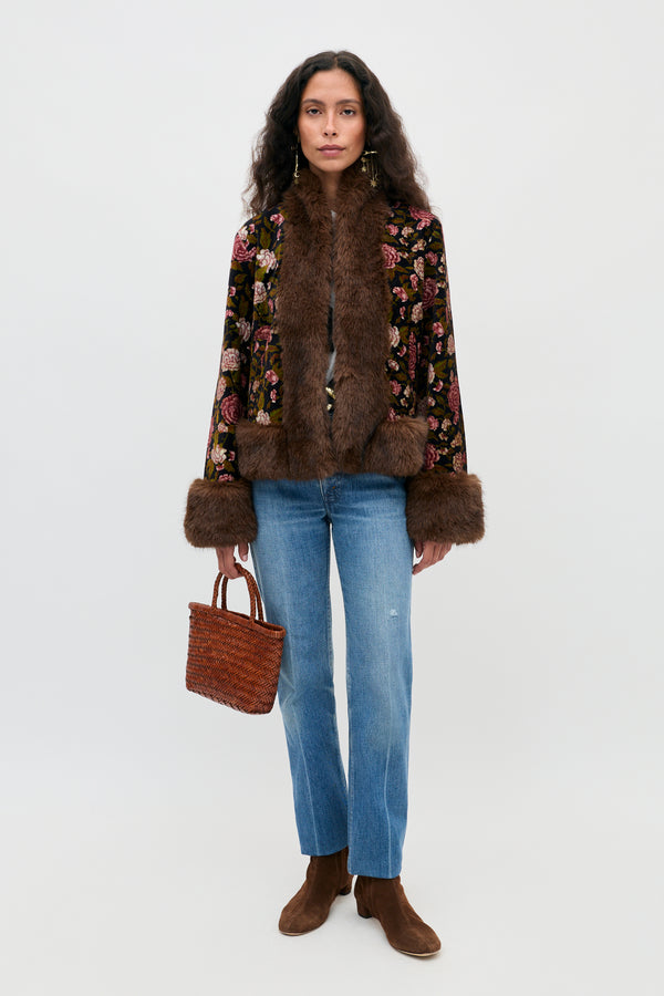 A woman stands facing forward, wearing the Jorgia - Enchanted Flora Green jacket with brown fur trim, blue jeans, brown boots, and holding a woven brown handbag. She has long wavy hair and a neutral expression against a plain background.