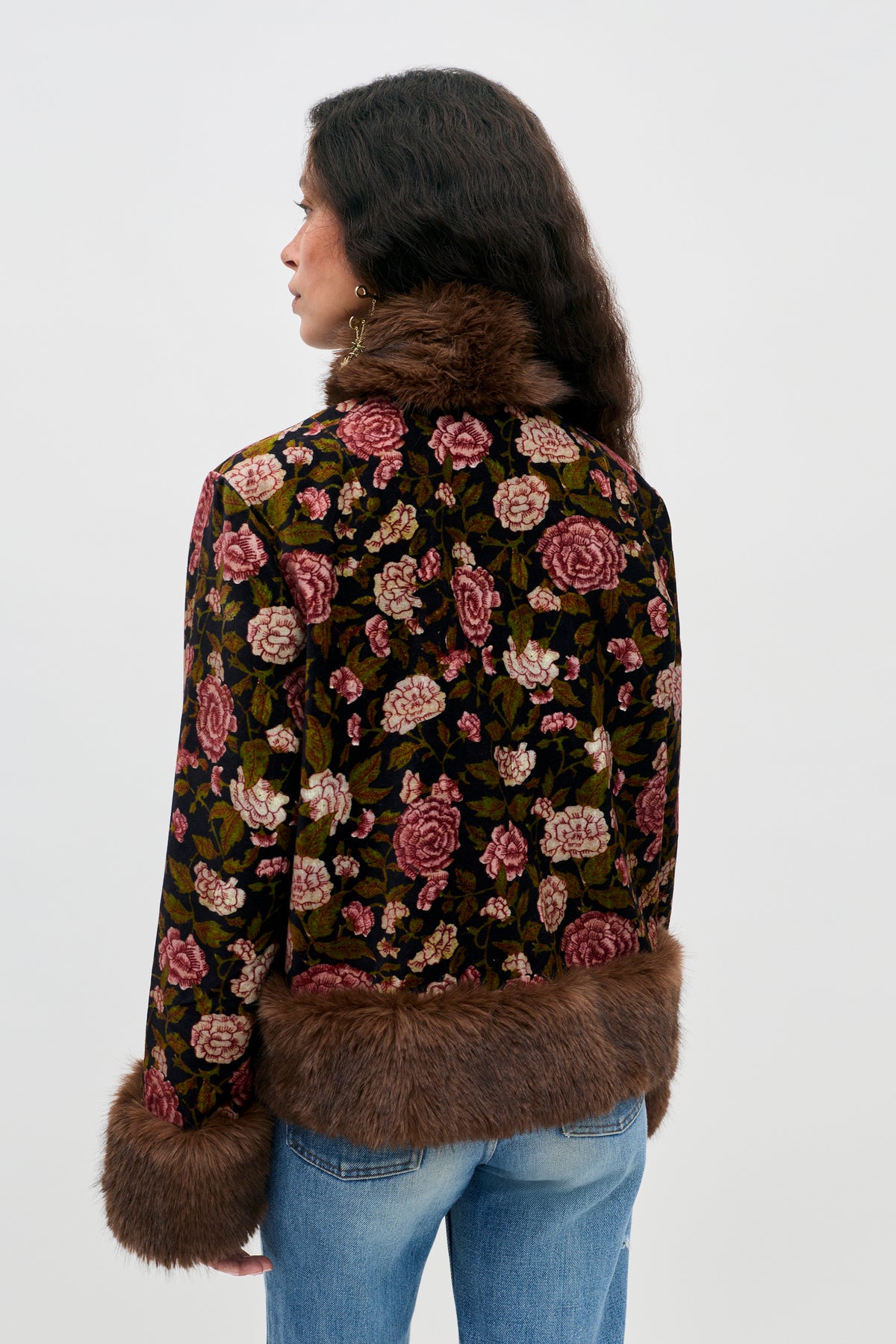 A person stands facing away, wearing the Jorgia - Enchanted Flora Green jacket, which features a floral pattern and faux fur trim on the collar, cuffs, and hem. They have wavy dark hair and are paired with blue jeans against a plain, light-colored background.