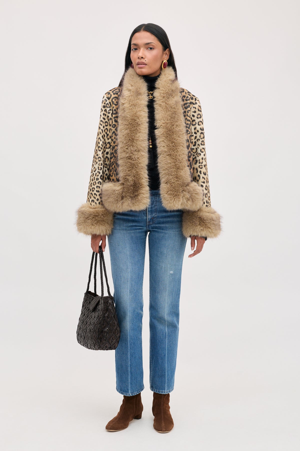 A model wearing a Jorgia - Savannah Leopard Brown jacket with faux fur trim, a black turtleneck, blue jeans, and brown suede ankle boots, carrying a dark woven handbag.
