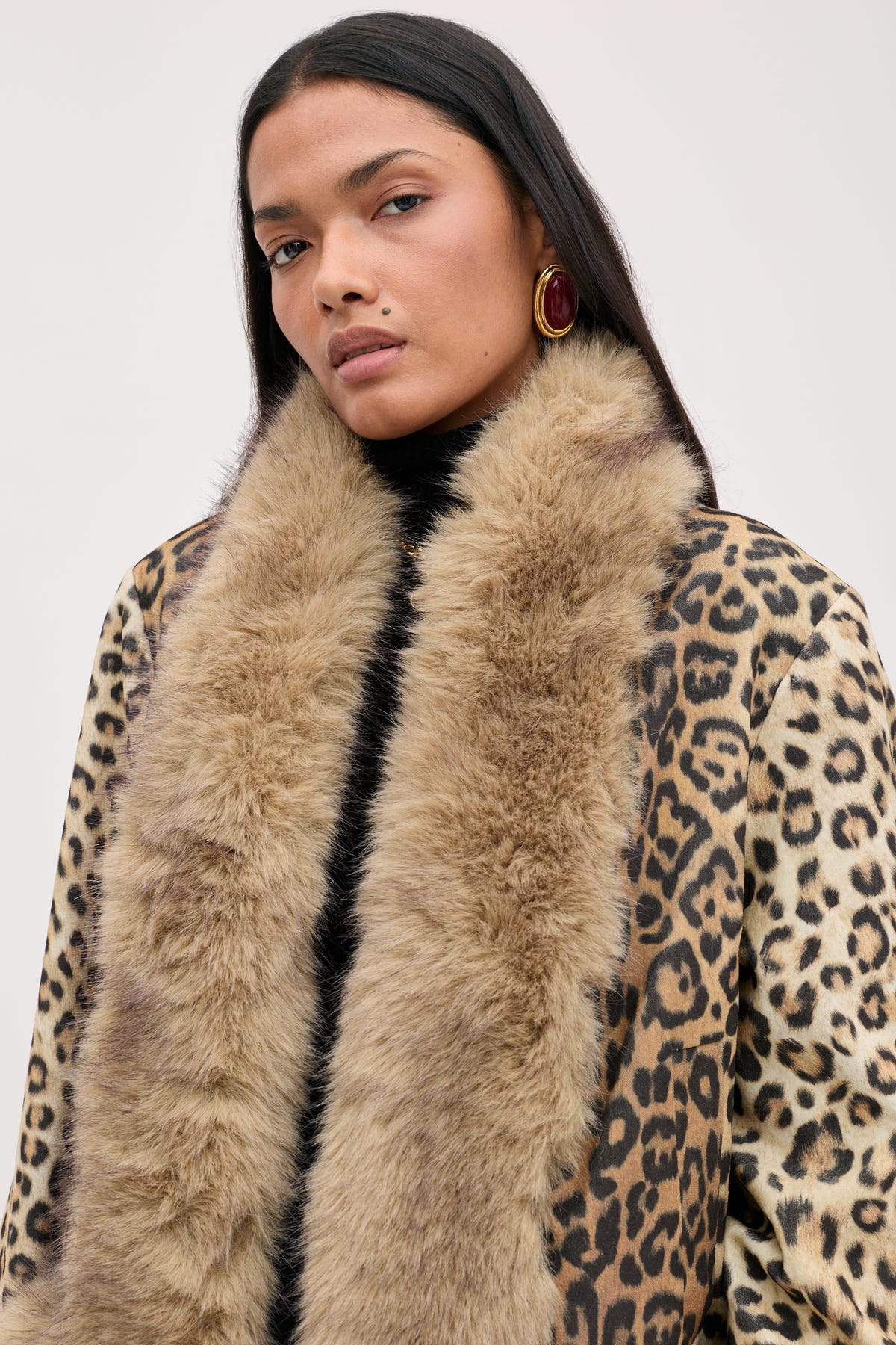 A person wearing a black turtleneck under a Jorgia - Savannah Leopard Brown jacket with a wide tan faux fur collar and large red earrings.
