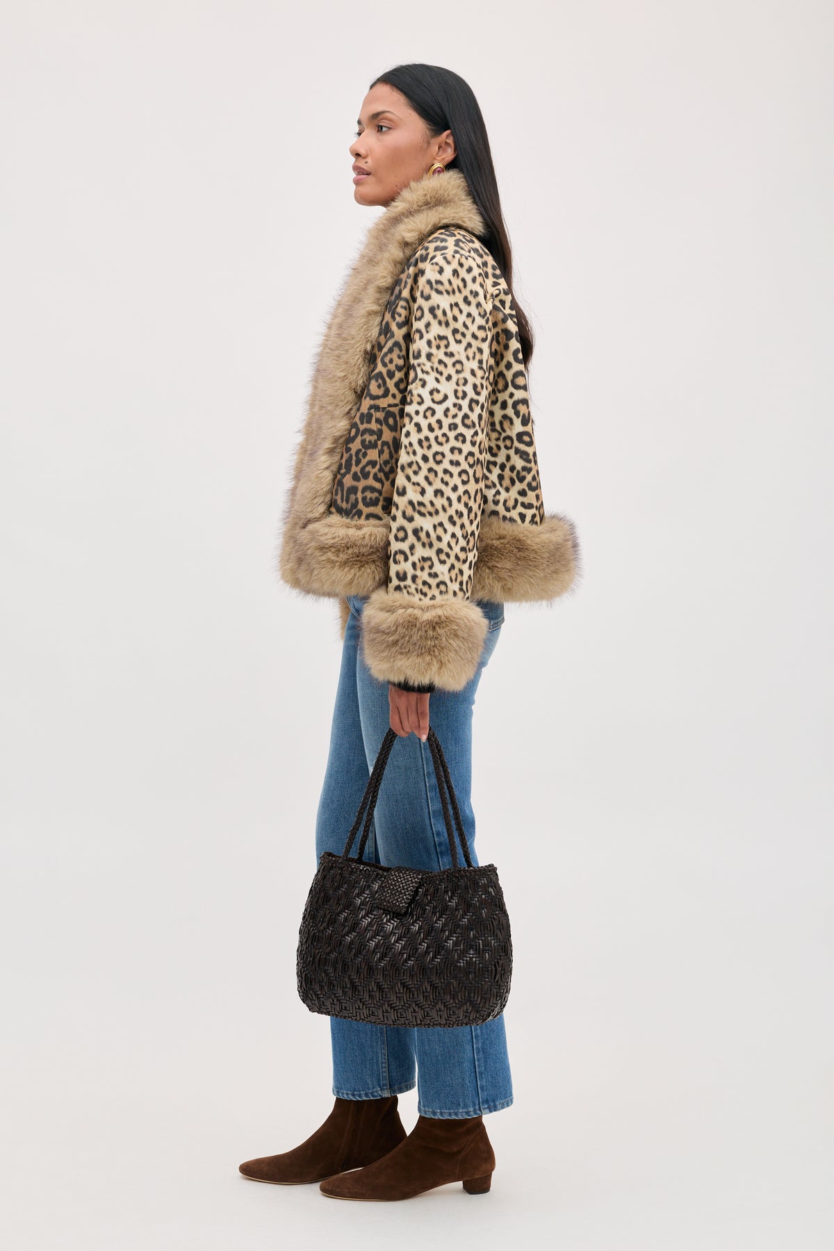 A person in profile wears a Jorgia - Savannah Leopard Brown jacket with fur trim, blue jeans, and brown boots, holding a dark woven handbag.