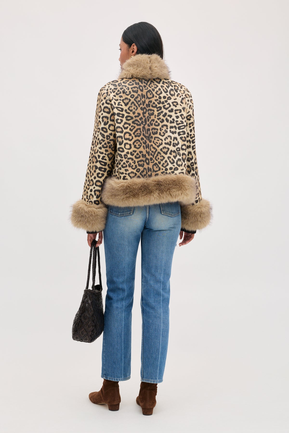 A person seen from behind wears the Jorgia - Savannah Leopard Brown jacket with fur trim at the collar, hem, and cuffs, paired with blue jeans, brown ankle boots, and a woven dark handbag.