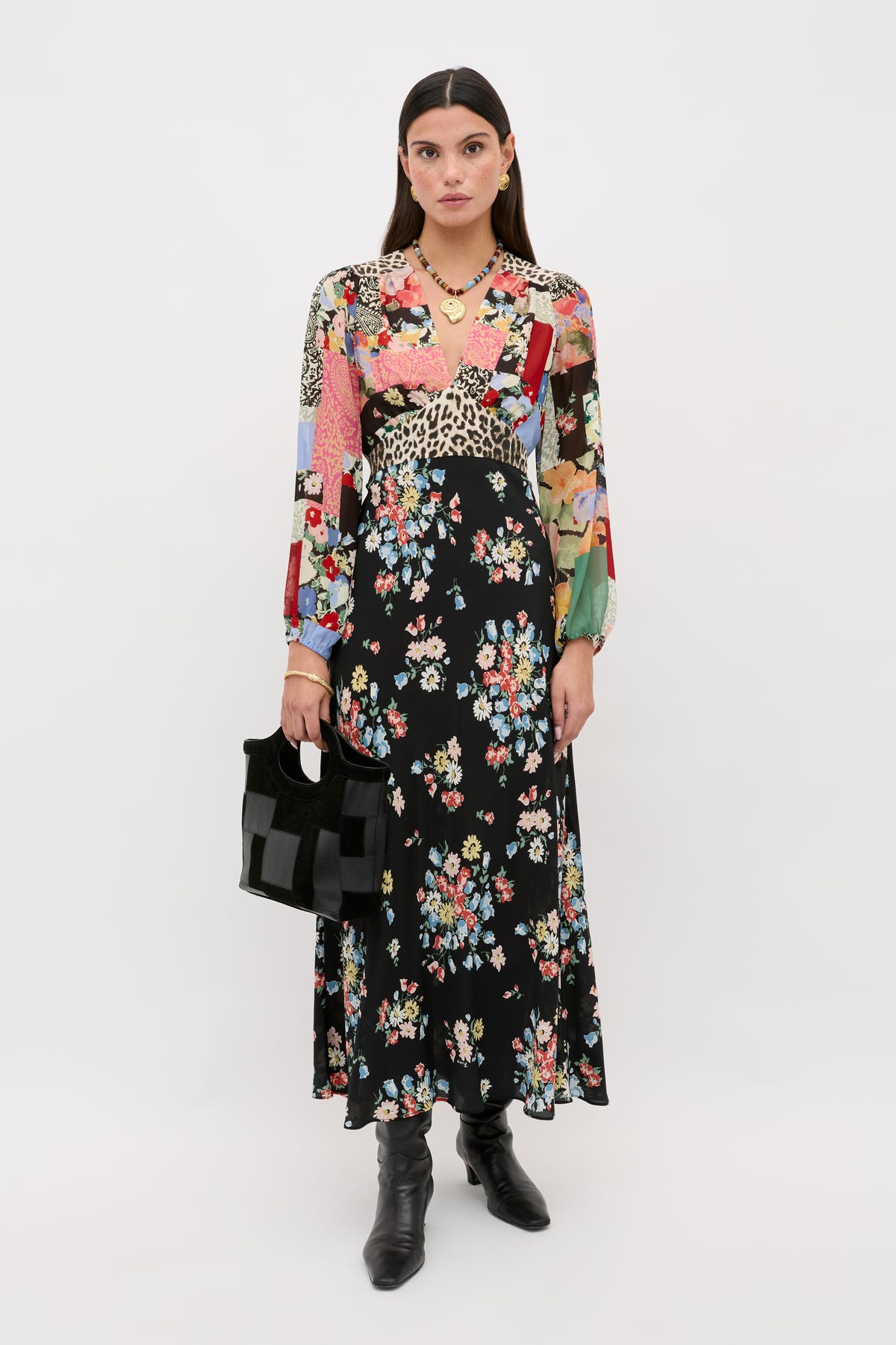 A woman wears a Kaila - Amelia Patchwork Mix long-sleeved, v-neck maxi dress with a multicolored patchwork bodice and a black floral skirt, paired with black leather boots and a black handbag.