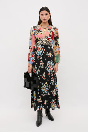 A woman wears a Kaila - Amelia Patchwork Mix long-sleeved, v-neck maxi dress with a multicolored patchwork bodice and a black floral skirt, paired with black leather boots and a black handbag.