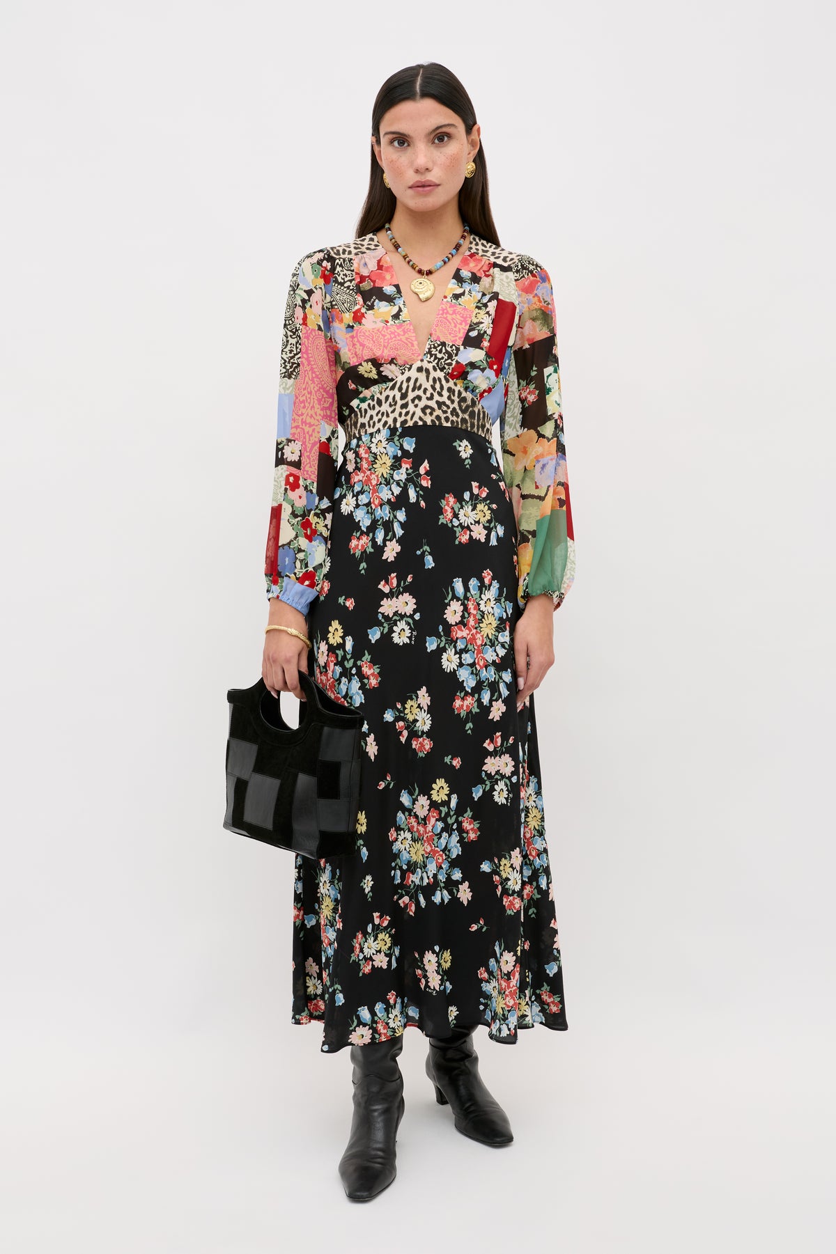 A woman models the Kaila - Amelia Patchwork Mix, a long-sleeved midi dress with a patchwork bodice and black floral skirt, styled with black boots and a matching handbag.