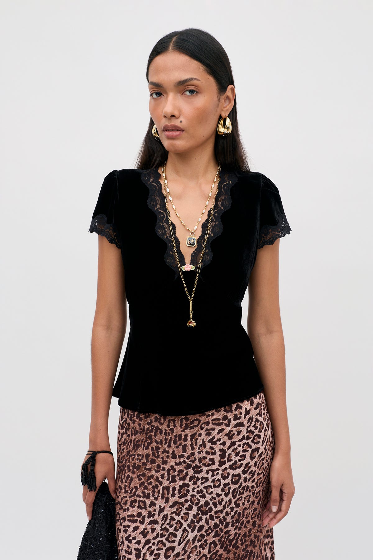 A model wearing the Karia - Black Velvet top with short sleeves, a lace-trimmed V-neckline, layered gold necklaces, and a leopard print skirt.