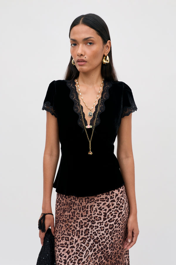 A model wearing the Karia - Black Velvet top with short sleeves, a lace-trimmed V-neckline, layered gold necklaces, and a leopard print skirt.