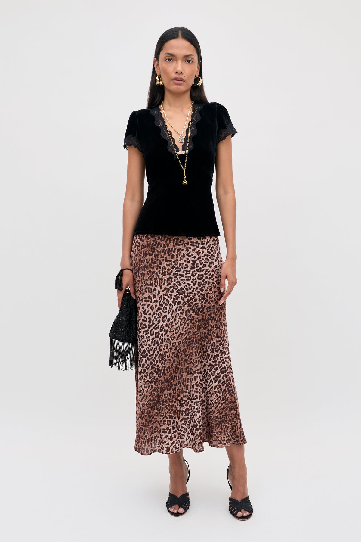 A woman wearing a Karia - Black Velvet top with lace trim, a leopard print midi skirt, and black heeled sandals, holding a small black fringed handbag.
