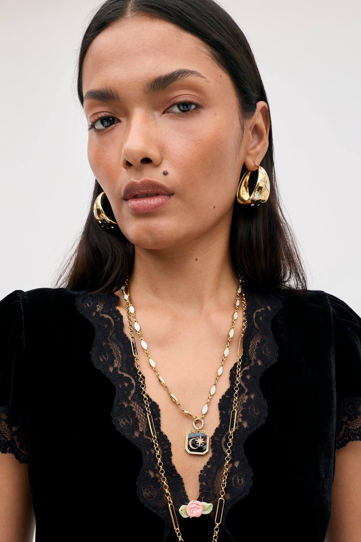 A model wears the Karia - Black Velvet top with lace trim, styled with gold hoop earrings and layered gold necklaces, including one with a square black pendant featuring a star and moon.