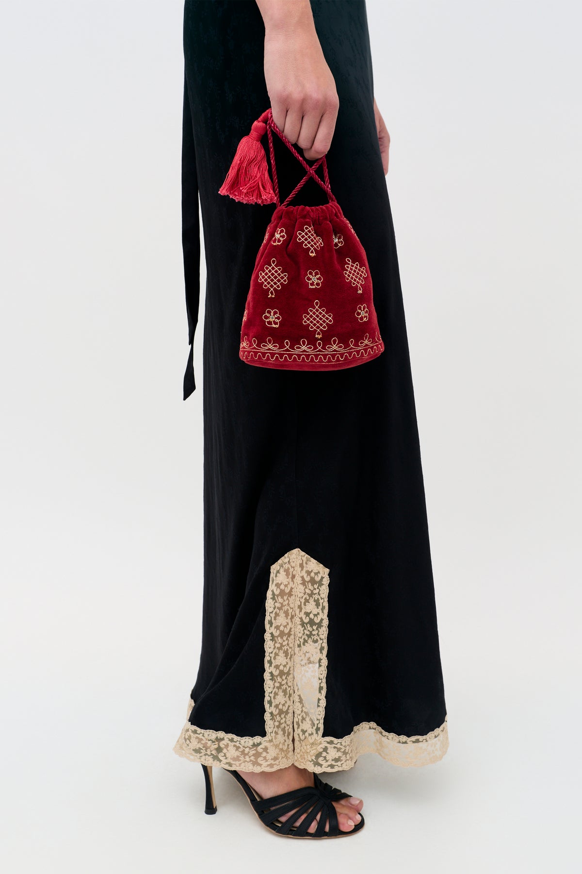 A person wearing an Orla - Waffle Soutache Embroidery Burgundy dress holds a small, red velvet drawstring pouch featuring gold embroidered patterns and a tassel.