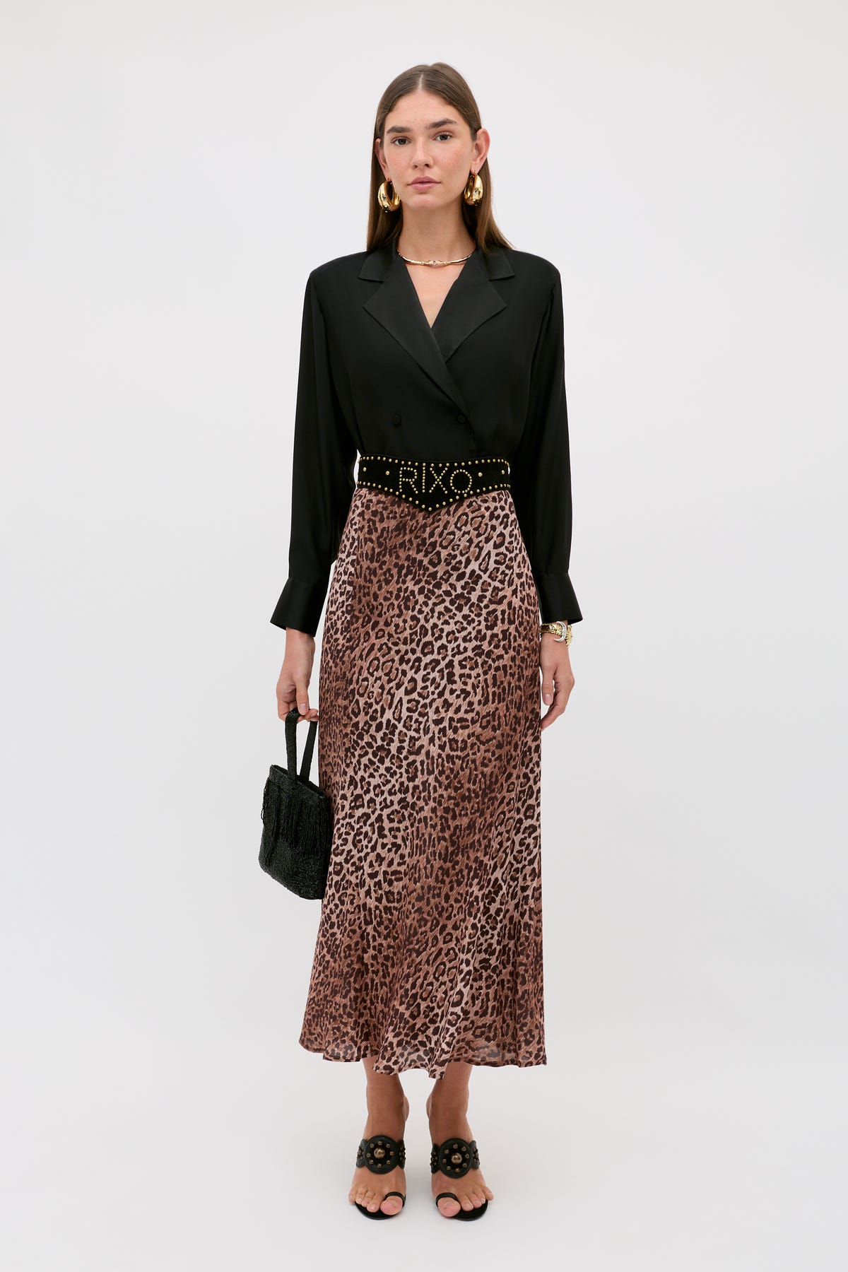 A woman stands against a plain background wearing the Kelly - Leopard Brown bias cut skirt in silk crepe de chine, styled with a black blouse, black belted waist, black sandals, gold earrings, and holding a black woven handbag.