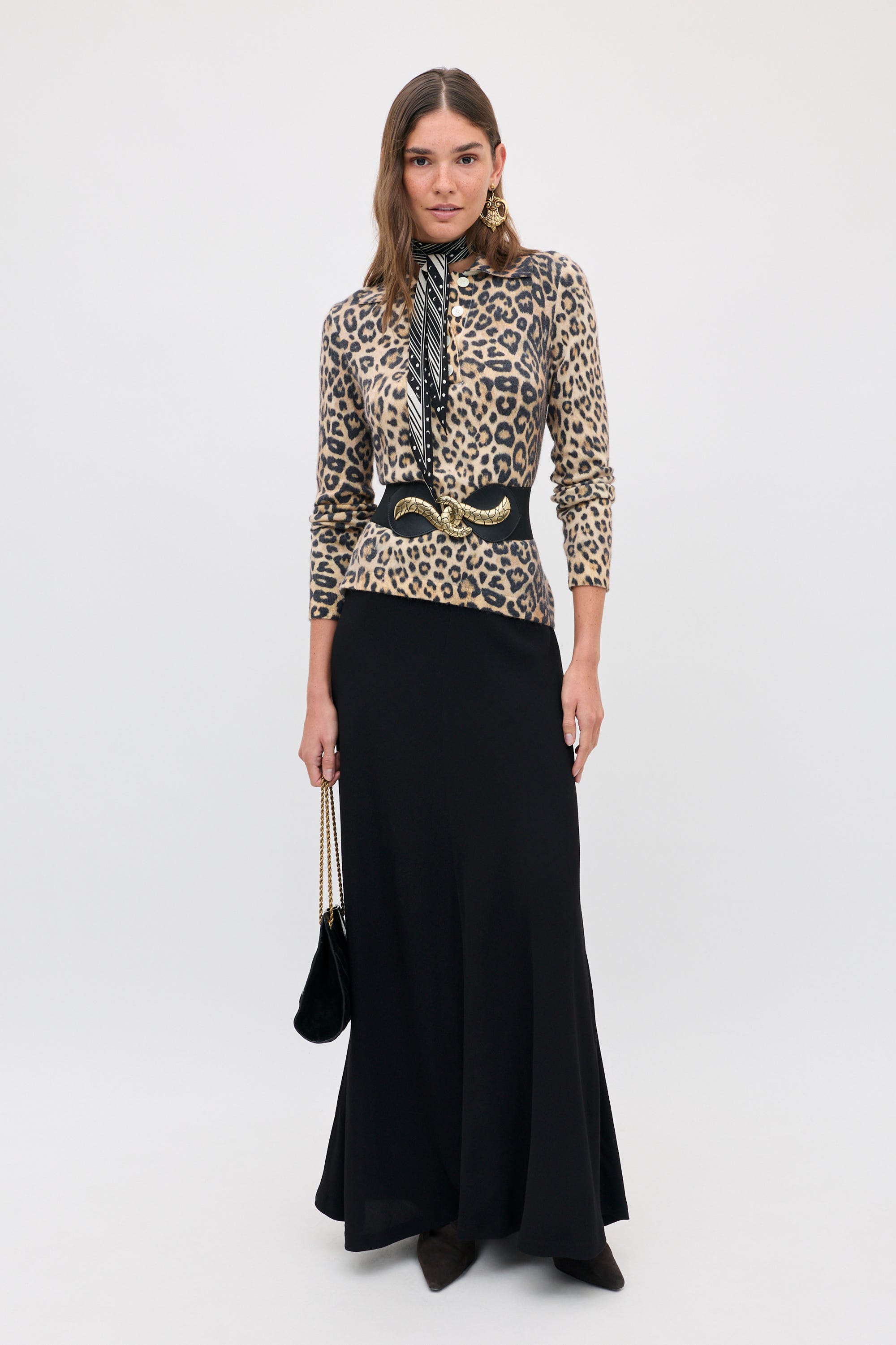 A woman wearing a Kristy - Savannah Leopard Brown long-sleeved polo shirt, a black waist belt with a gold buckle, a patterned neck scarf, and a long black skirt, holding a small black shoulder bag.