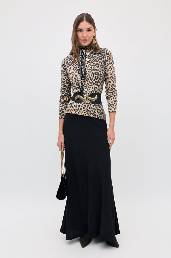 A woman wearing a Kristy - Savannah Leopard Brown long-sleeved polo shirt, a black waist belt with a gold buckle, a patterned neck scarf, and a long black skirt, holding a small black shoulder bag.