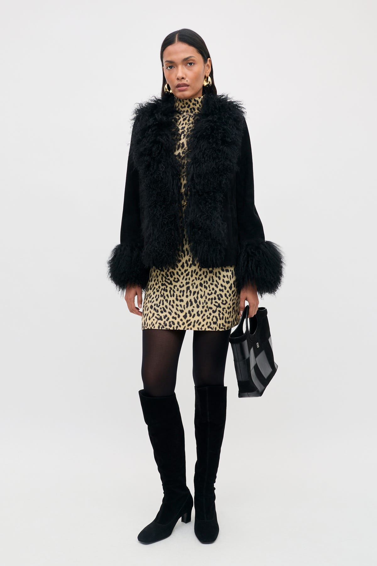 A person wearing a Sammy - Black jacket with fur trim over a leopard-print dress, black tights, tall boots, and holding a black handbag.
