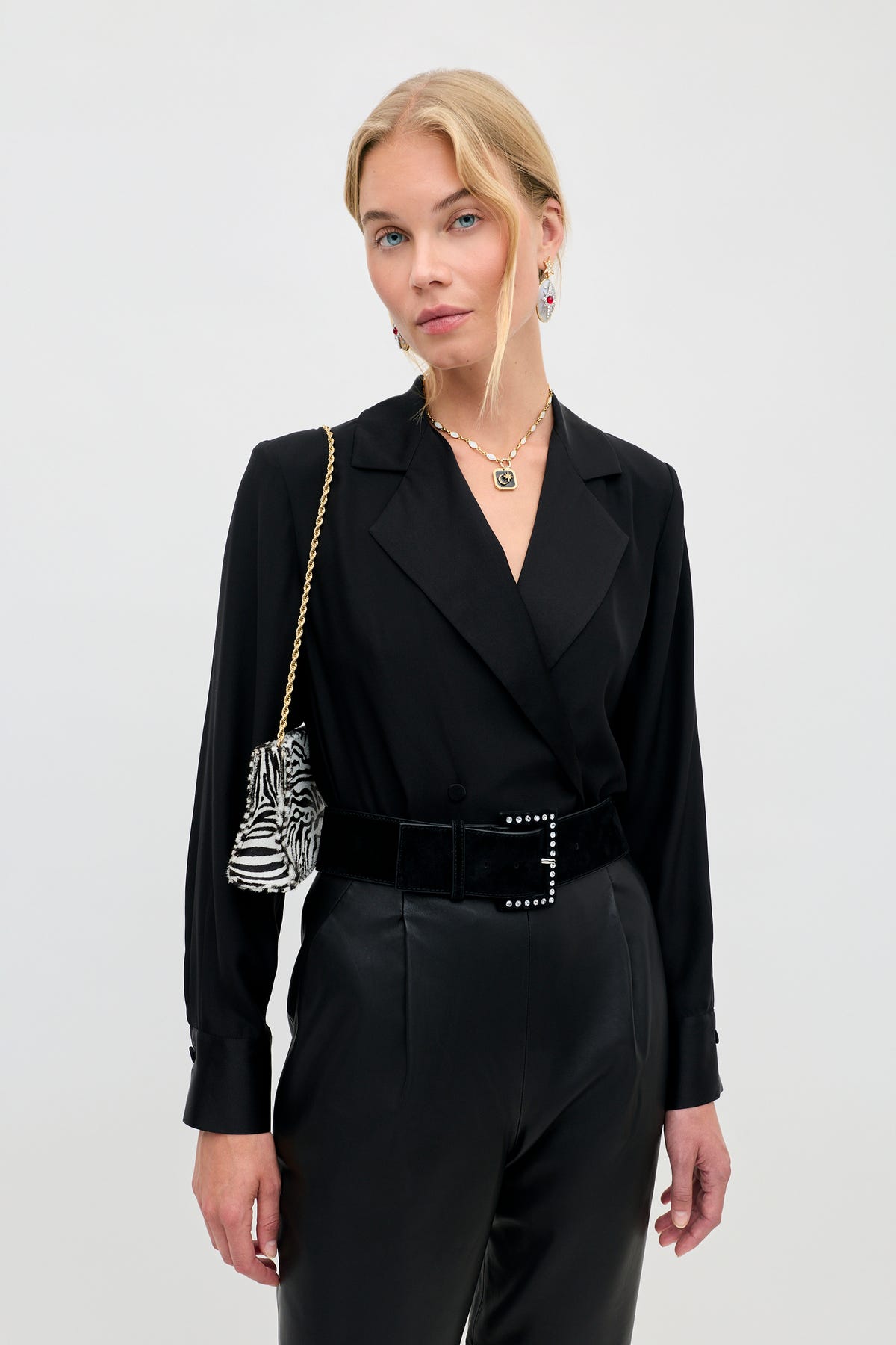 A woman wears a Lorraine - Black long-sleeve blazer with a wide belt and a small zebra-print handbag on a gold chain.
