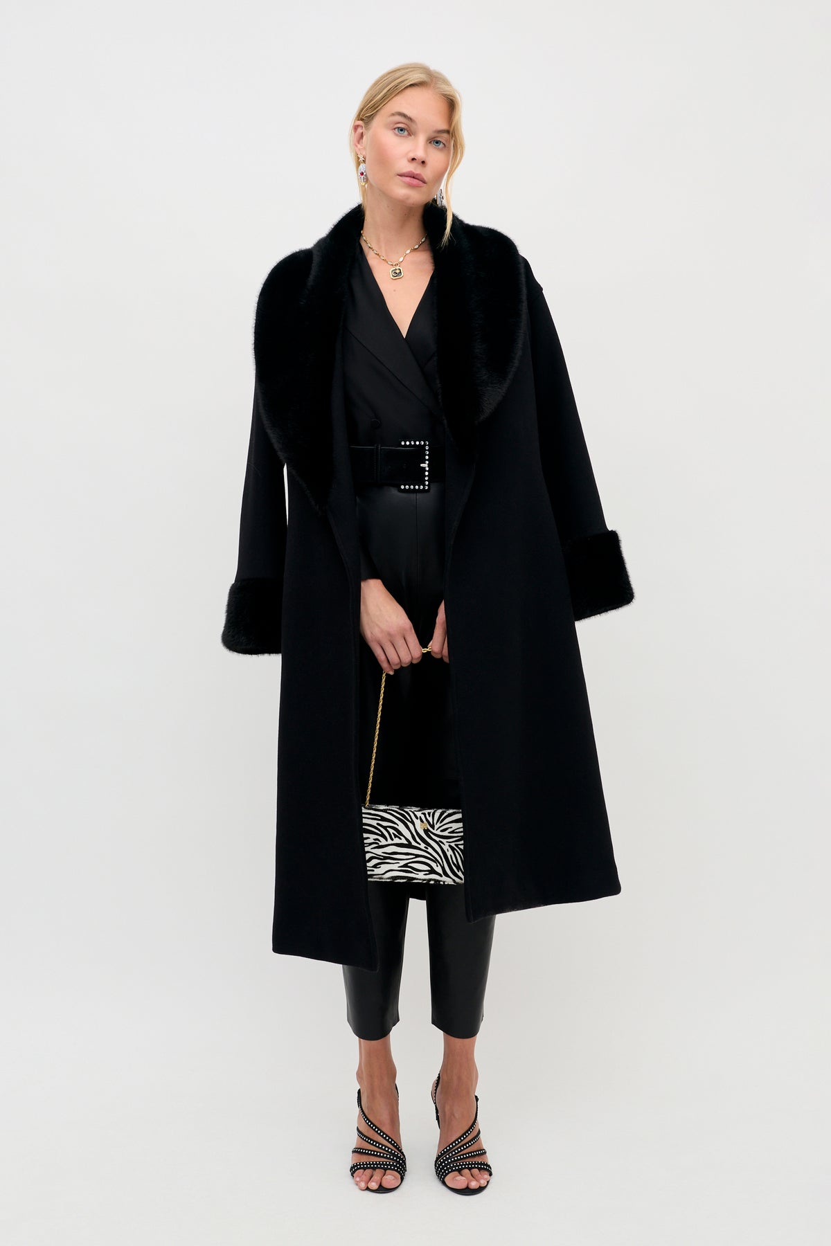 A woman wearing a Lorraine - Black coat with a faux fur collar and cuffs over a black belted outfit, holding a zebra-print handbag.