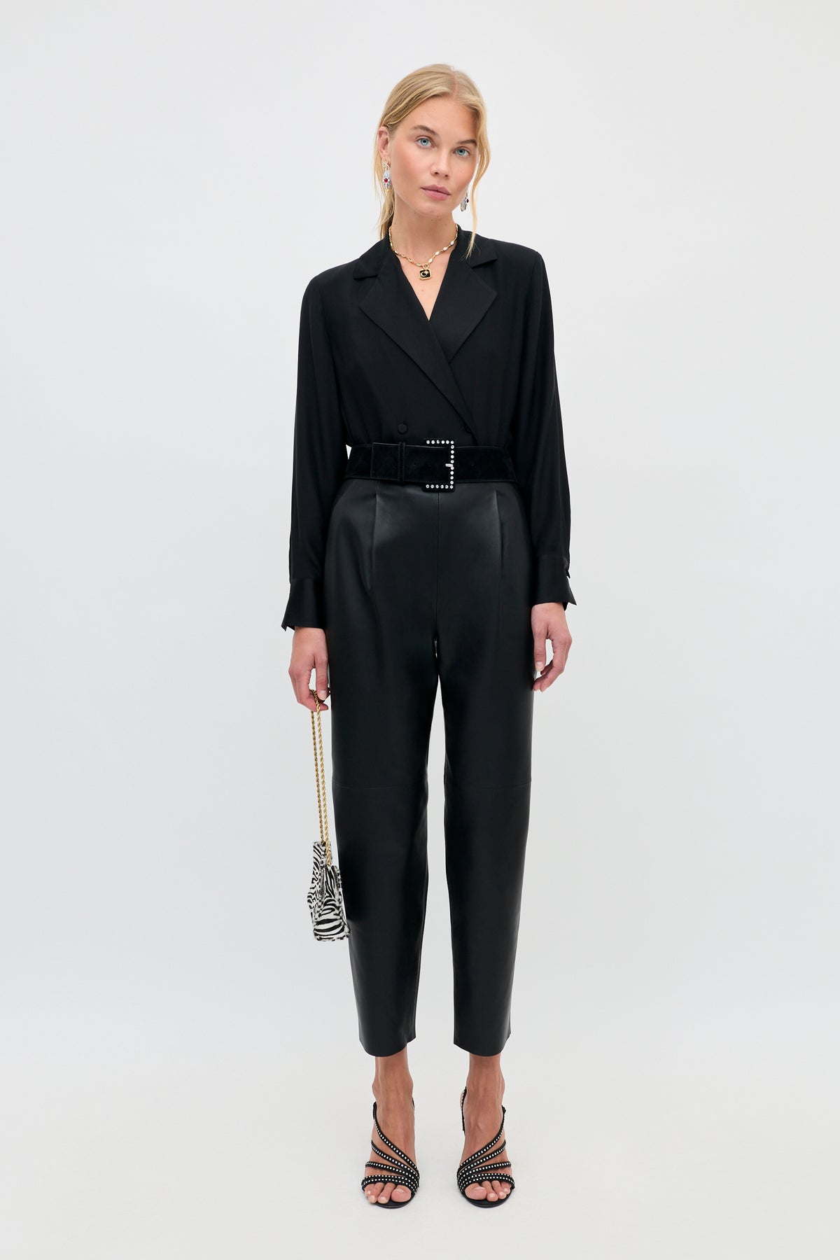 A woman wearing a Lorraine - Black long-sleeved button-down top tucked into black trousers with a wide buckled belt, carrying a small zebra-print bag and wearing strappy heeled sandals.