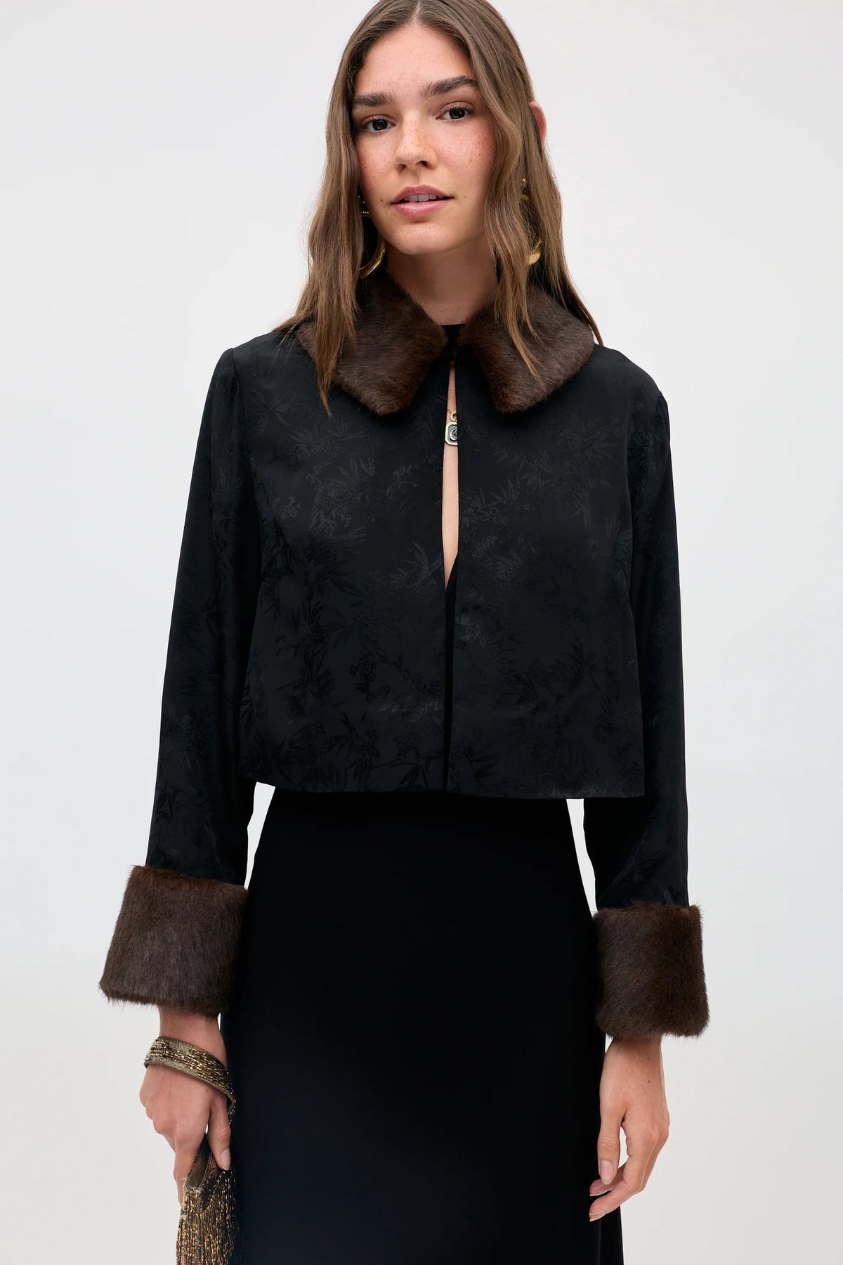 A woman wearing the Luanne - Black jacket, featuring a subtle floral pattern, brown faux fur collar and cuffs, paired with a black skirt, stands against a light background.