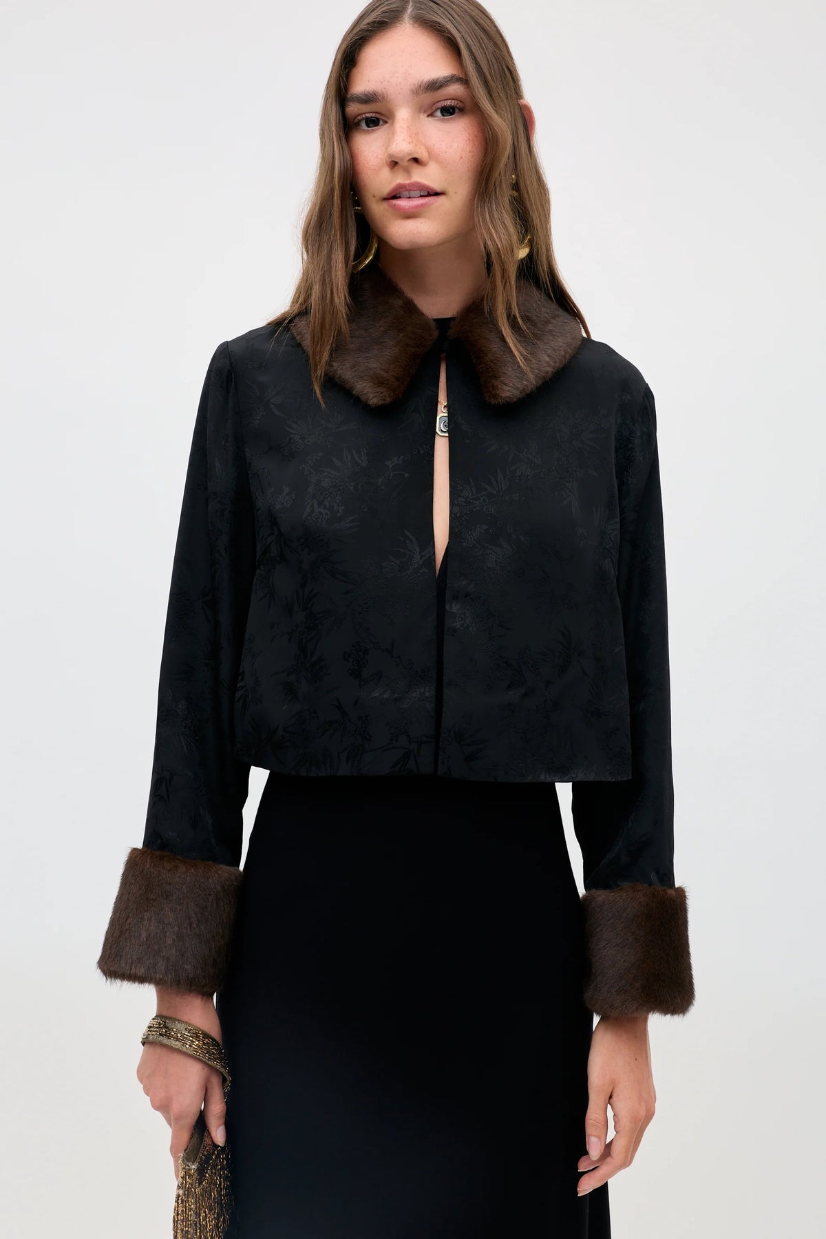 A woman wearing the Luanne - Black jacket, featuring a subtle floral pattern, brown faux fur collar and cuffs, paired with a black skirt, stands against a light background.